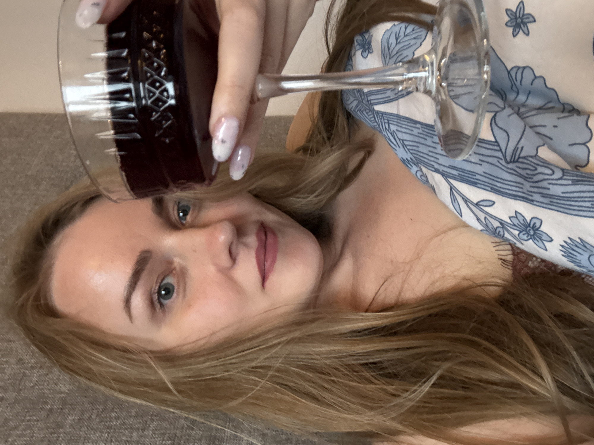 A young woman with blonde hair is holding a glass of red wine. She has light skin and blue eyes. Her nails are painted with a light color and have silver sparkles. She is wearing a patterned shirt or dress with a blue and white floral design. The background is a plain, grey surface. The location is unknown.