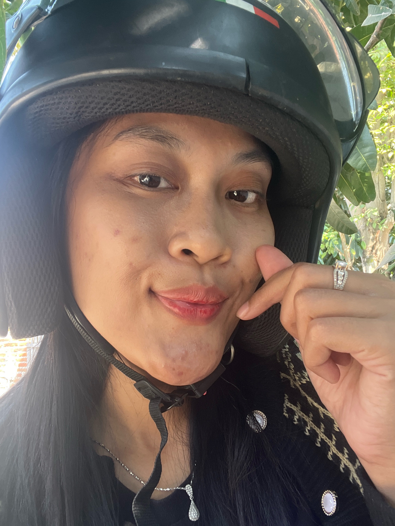 A close-up selfie shows a woman wearing a black helmet, with some of her face and chest visible. She has dark hair and is wearing a black top with a patterned design and silver-toned buttons. A silver necklace with a pendant is visible around her neck. She is also wearing a ring on her right hand. The background appears to be an outdoor setting with greenery and a hint of brickwork.