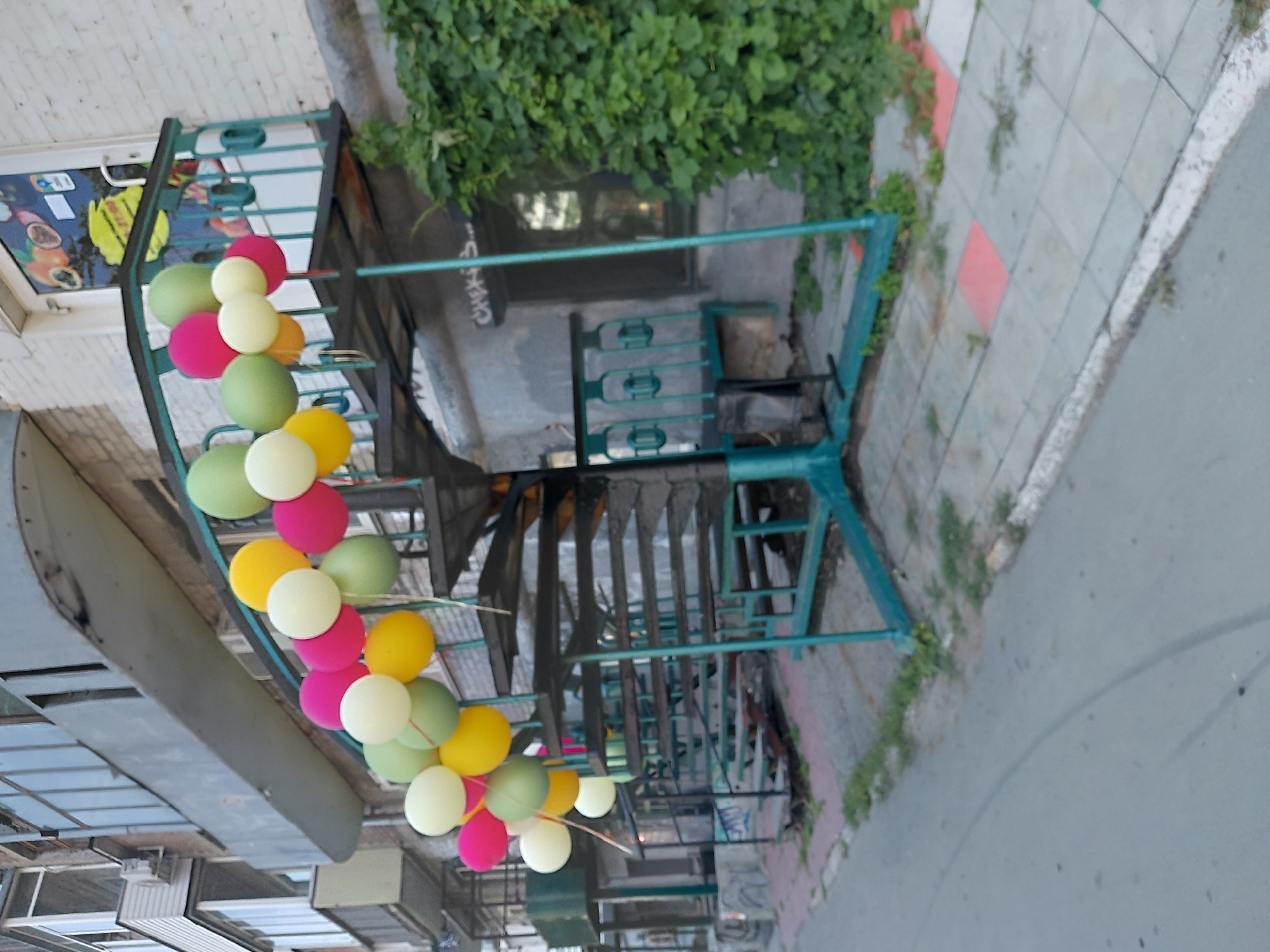 An outdoor spiral staircase with a teal-colored metal frame, decorated with a string of colorful balloons, leads up to a balcony of a multi-story building. The balloons are in shades of green, yellow, pink, and white. The building's facade is made of light-colored brick. Green foliage grows on the right side of the image, adjacent to the building. A sidewalk with a patterned tile surface is visible in the foreground, and a street runs alongside it. There are no people or animals visible in the image. The scene appears to be in an urban environment.