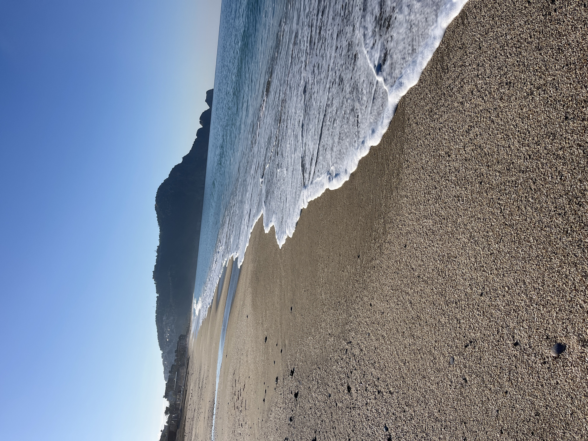 The image captures a beach scene, likely in Alanya, Turkey, given the mountainous terrain and coastal setting. The sky is clear blue transitioning to a lighter hue near the horizon. A dark, rugged mountain ridge is visible, partially obscured by haze. The sea is characterized by white foamy waves crashing onto a coarse, sandy beach. The sand is dry, with a gradient of light brown to dark brown, and interspersed with pebbles. There are no visible people or objects on the beach, besides natural elements. The scene conveys a sense of tranquility and natural beauty.