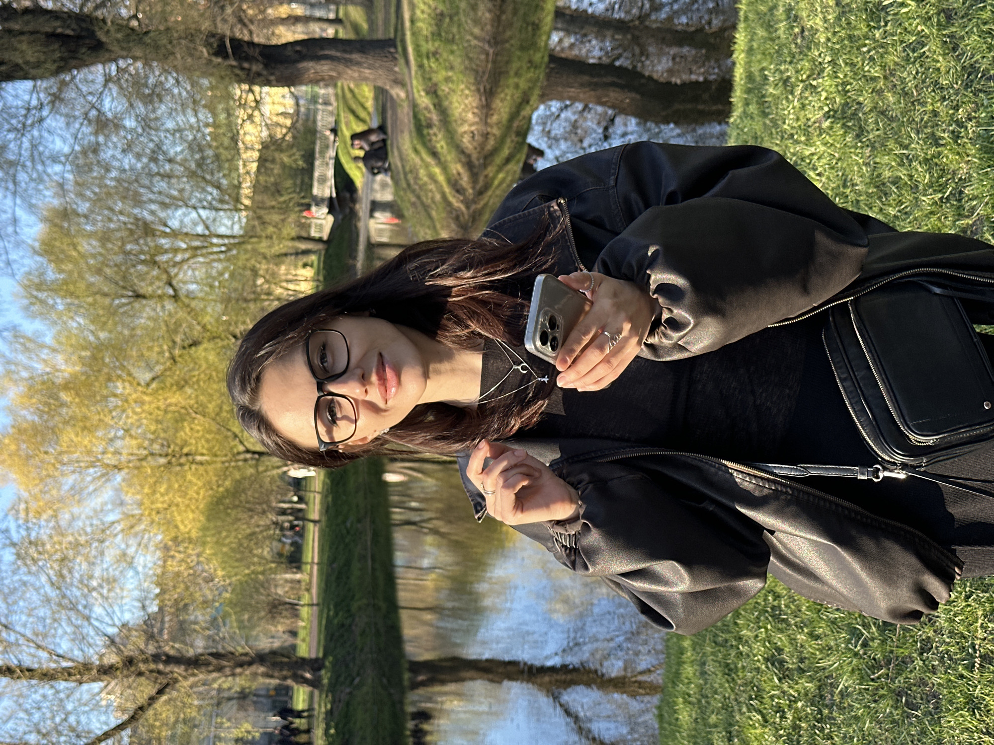 The image depicts a woman in a black leather jacket, black top, and glasses, holding a smartphone. She stands outdoors on green grass, near a body of water that reflects trees and sky. The scene appears to be a park or garden within the city of Saint Petersburg, Russia, as evidenced by the presence of trees, water, and visible urban elements in the background. The lighting suggests it is daytime, with dappled sunlight across the grass. Other details are that she has a silver necklace on. A black purse is strapped over her shoulder. She has rings on her left hand.