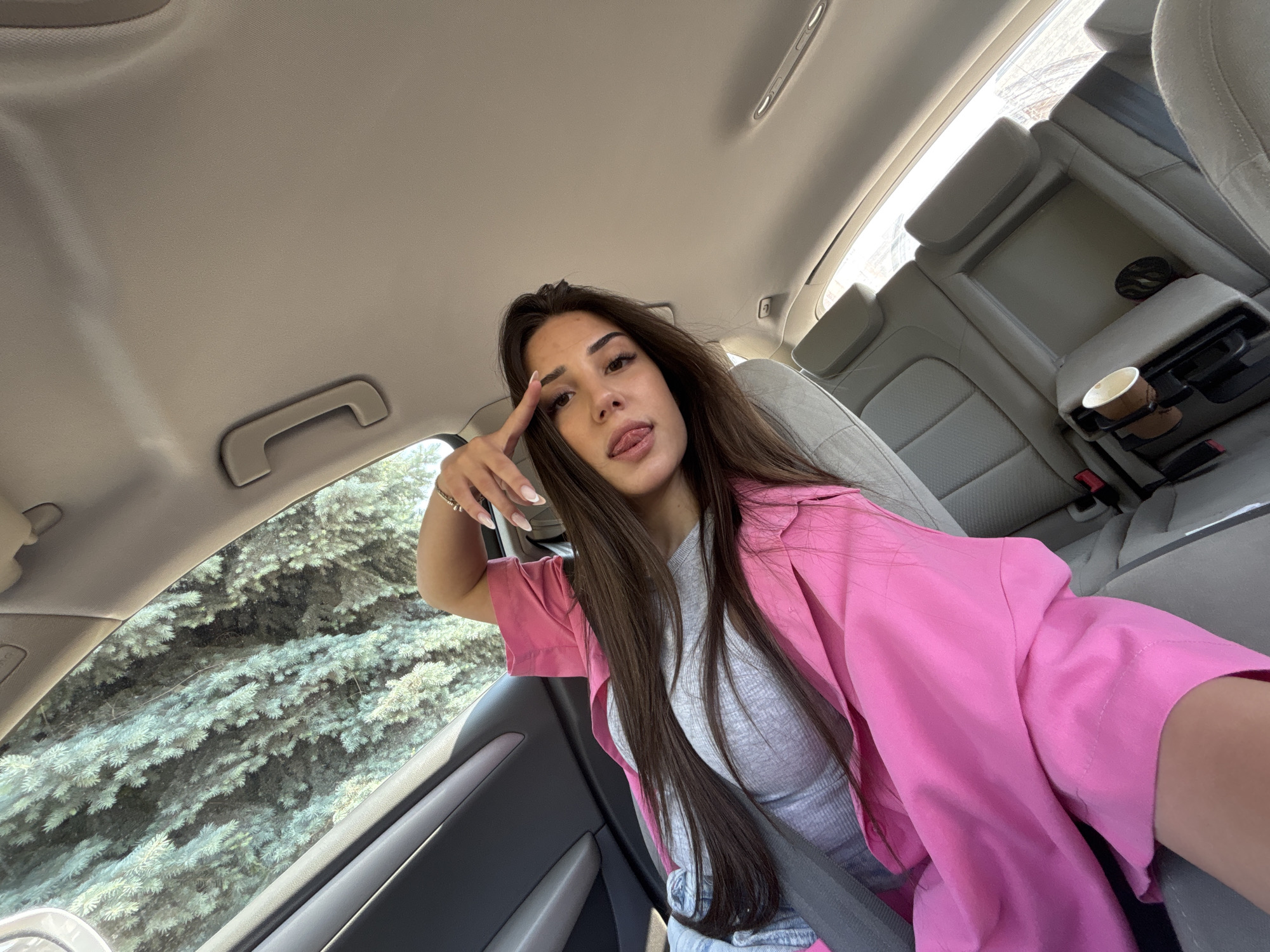 A young woman takes a playful selfie inside a car, likely in Plovdiv, Bulgaria, during bright daylight hours. She is positioned in what appears to be the driver's or front passenger's seat, leaning towards the camera with her long, dark brown hair flowing over her shoulders. She wears a grey ribbed tank top beneath a vibrant pink button-down shirt or lightweight jacket. Her right hand is raised to her temple, forming a peace sign, and she is sticking her tongue out slightly, conveying a cheerful and confident mood. Her nails are long and feature a French tip design, and a thin bracelet adorns her right wrist.
The car's interior is visible, featuring light grey/beige upholstery on the seats and headliner. A grab handle is present on the passenger side roof. Through the left car window, dense green foliage, possibly evergreen trees, can be seen, indicating the vehicle is outdoors, perhaps parked by a roadside or in a green area. The bright lighting suggests a sunny day. In the background, the rear seats are visible, with a central armrest folded down to reveal cup holders containing a light brown paper cup. No visible text is present in the image.