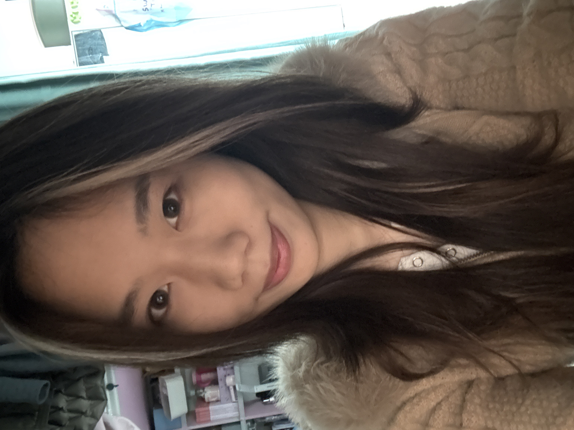 The scene is an indoor setting within the city of Nan'an District, China. A single individual, appearing to be a female of East Asian ethnicity in her twenties or early thirties, is positioned centrally, facing directly towards the camera. She has long, dark brown hair with a prominent light-colored streak on the viewer's left side, framing her face. Her eyes are dark brown, and she displays a slight, closed-lip smile. She is wearing a light beige or tan textured outerwear, possibly fleece or faux fur, over a white inner garment with a visible silver snap button and a portion of a zipper closure. The background is out of focus; on the viewer's left, a light pink multi-tiered shelving unit is visible, holding various small containers and bottles. On the viewer's right, a bright window or light source is partially obscured by a teal curtain, with indistinct forms and colors visible on a surface behind it. The lighting is soft, illuminating the subject's face.