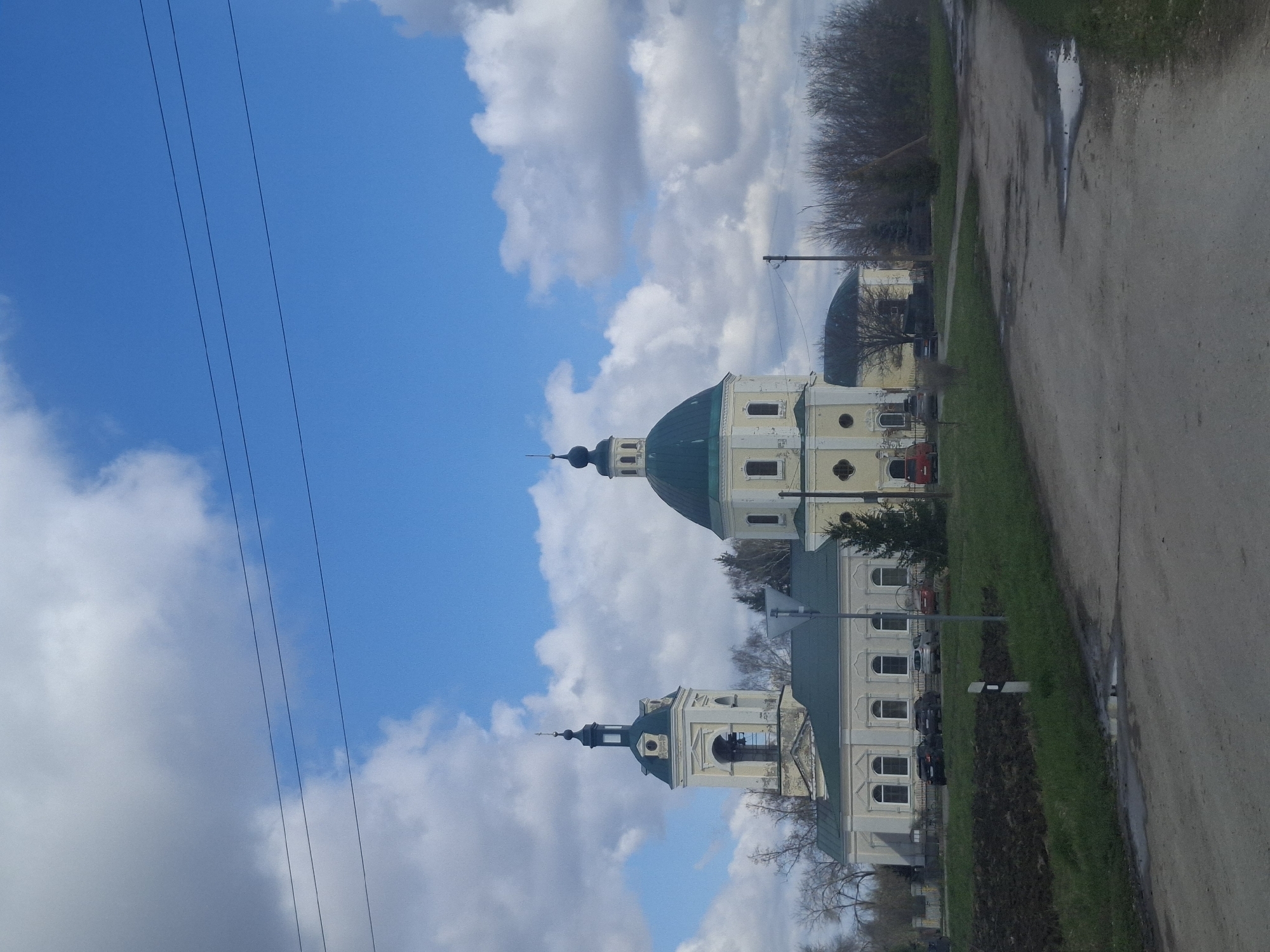 This image presents a serene daytime landscape, likely set in Паниковец, Russia, characterized by its architectural and natural elements. The main subjects are two prominent religious buildings, resembling Orthodox churches or cathedrals, with light-colored (possibly cream or light yellow) facades and distinctive green domes and spires. The structure on the right features a large central dome and a smaller spire, while the one on the left has a bell tower topped with a spire. Both buildings exhibit multiple arched windows.
In the foreground and midground, a dirt or gravel road runs along the right side of the frame, showing a visible puddle that suggests recent rain. Green grass lines the edge of the road and extends further into the scene. Bare deciduous trees, indicating late autumn, winter, or early spring, are scattered around the buildings and along the landscape. A few vehicles are faintly discernible parked behind the left-most building.
The sky is a bright blue, adorned with large, fluffy white and grey clouds. Several power lines stretch across the upper left section of the image. No people are visible, contributing to the quiet and undisturbed atmosphere of the scene. There is no visible text in the image.