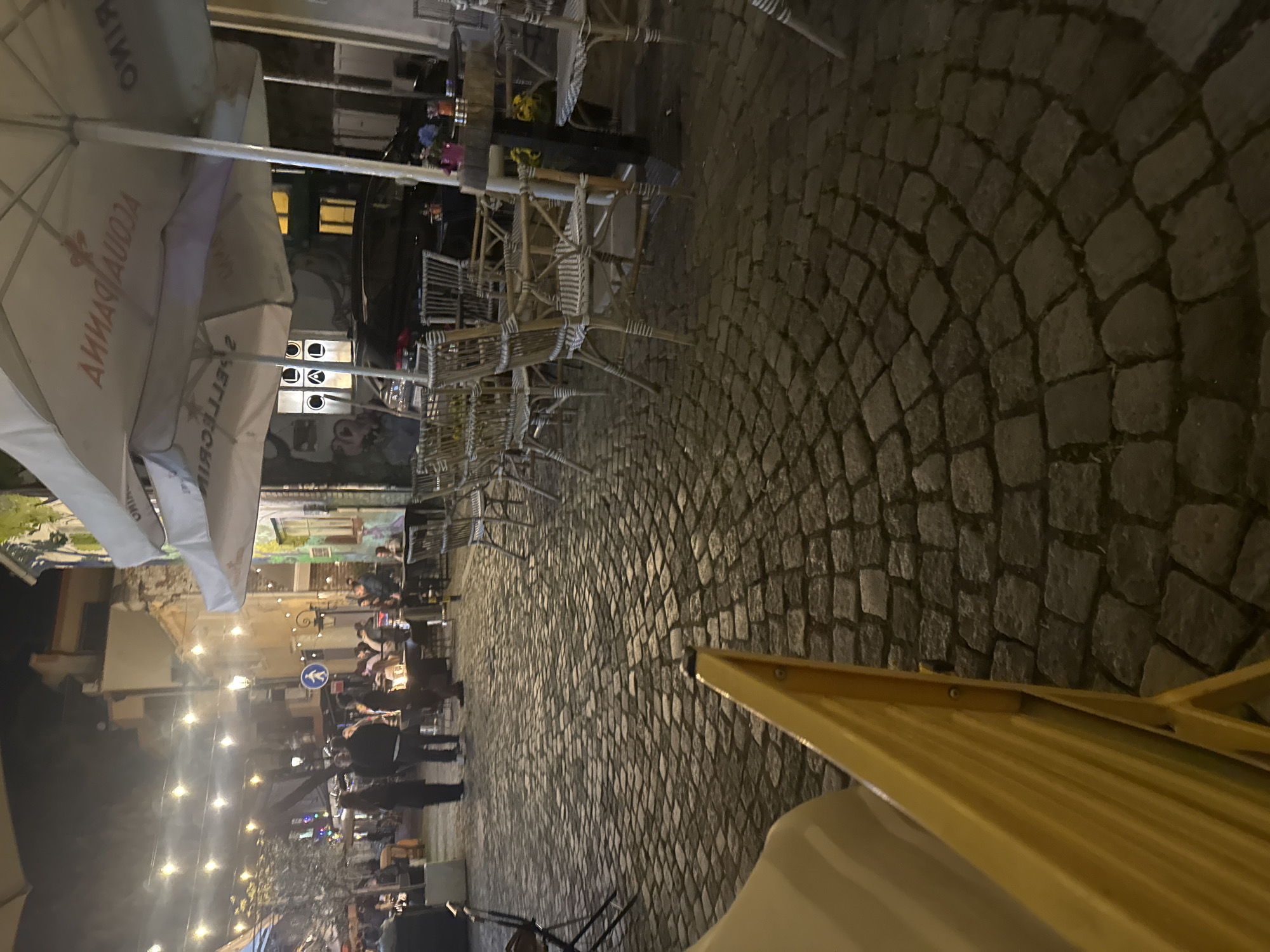 The image captures a street scene in Plovdiv, Bulgaria, at night. The foreground features a restaurant with tables and chairs along a cobblestone street. Tables are arranged under large sun umbrellas with branding visible. Pedestrians are walking along the street, and people are seated at the restaurant tables. The buildings along the street are lit, and streetlights and decorative lights illuminate the scene. The street curves away from the camera's perspective.