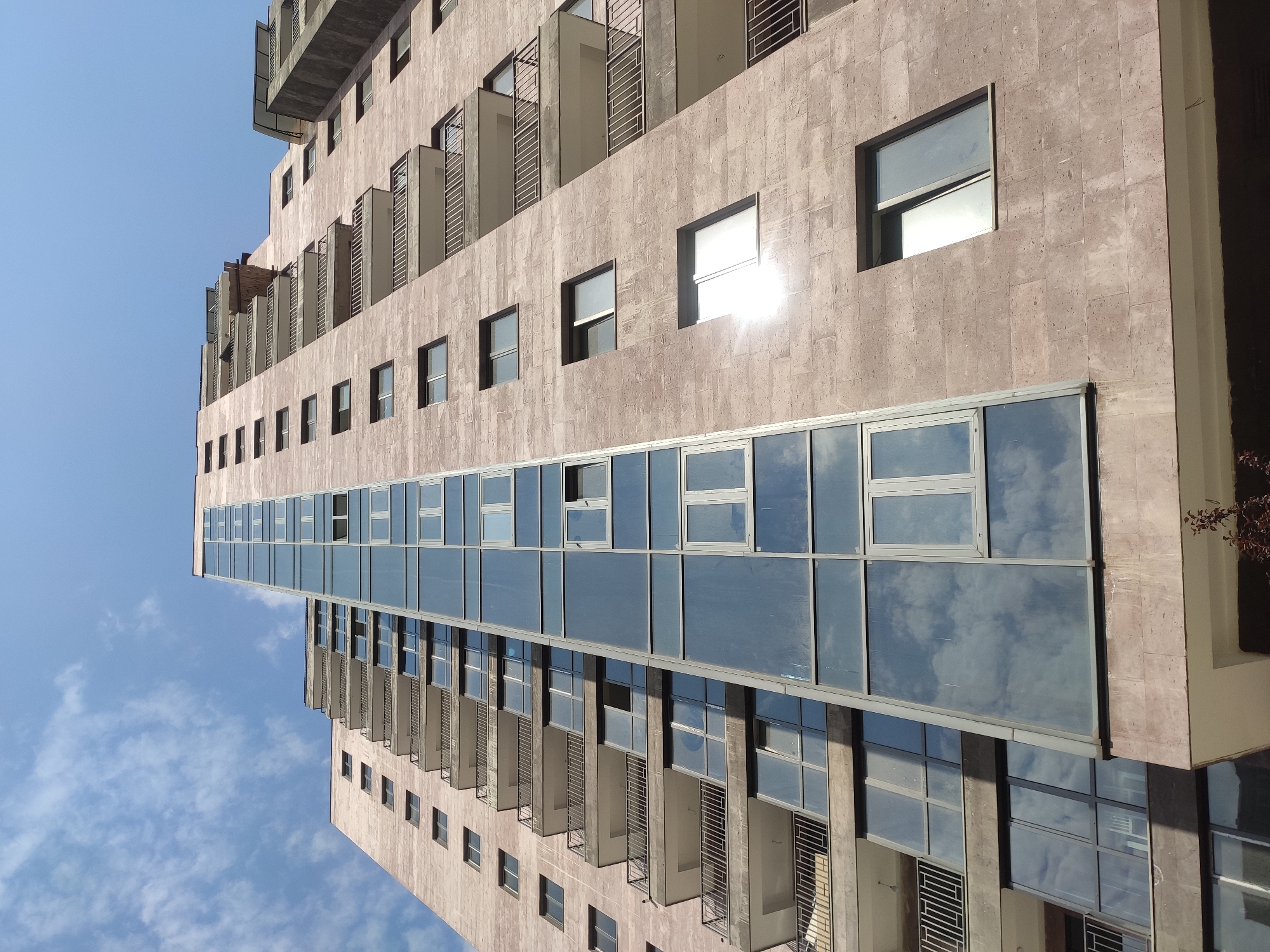 This is a low-angle, full shot of a modern, tall apartment building in Yerevan, Armenia, under a bright blue sky with scattered clouds. The building features a segmented design with sections clad in light pinkish-brown stone and a prominent, vertical glass facade that reflects the sky. The stone portions of the building have numerous balconies with metal railings, and regular, rectangular windows. The glass section is comprised of large, rectangular panes, some of which are open. The building appears to be residential.
