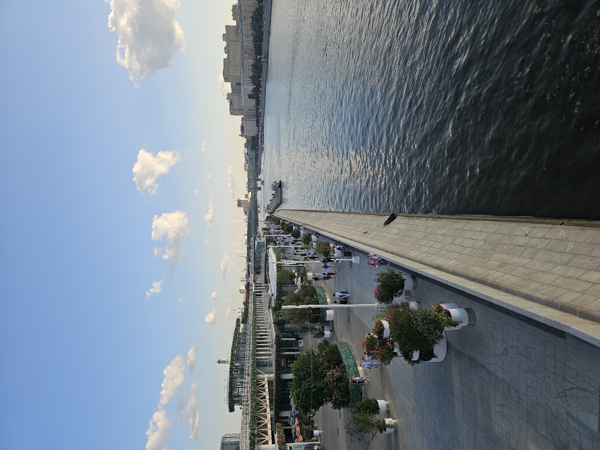 The image shows a wide, calm river in Moscow, Russia, with a pedestrian promenade along its left bank. The promenade is lined with planters containing flowers and shrubs, as well as white cylindrical pots. Several people are walking along the promenade. On the left side of the promenade is a large, modern building with multiple levels, covered in greenery and adorned with floral decorations. In the background, across the river, are several large buildings, characteristic of Moscow's architecture, and a bridge spanning the river. The sky is blue with scattered white clouds, indicating it is daytime. The scene appears to be a public space designed for leisure and recreation.