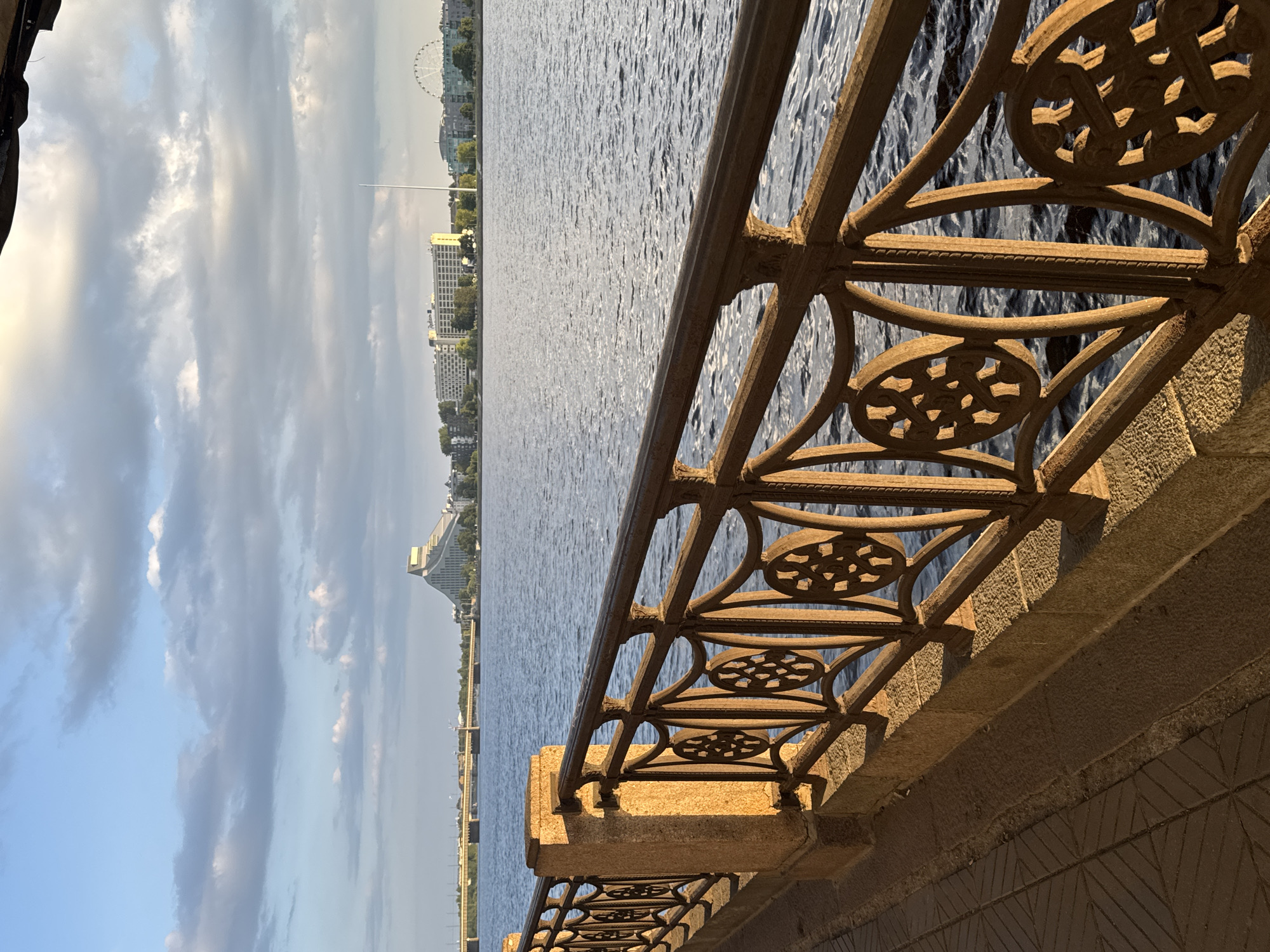 The image depicts a waterfront view in Riga, Latvia. In the foreground, a decorative metal railing with stone supports runs along a paved walkway. The railing has intricate circular patterns and ornate scrolling. Beyond the railing is a body of water, likely the Daugava River, with gentle ripples reflecting the light.
In the background, across the water, the cityscape of Riga is visible. Prominent structures include the distinctive, pyramid-shaped Latvian National Library, a large hotel building labeled "Radisson," and a Ferris wheel. A bridge spans the river in the distance. The sky above is partly cloudy, with sunlight casting a warm glow on the scene, particularly on the railing and walkway. There are no people visible in the image.