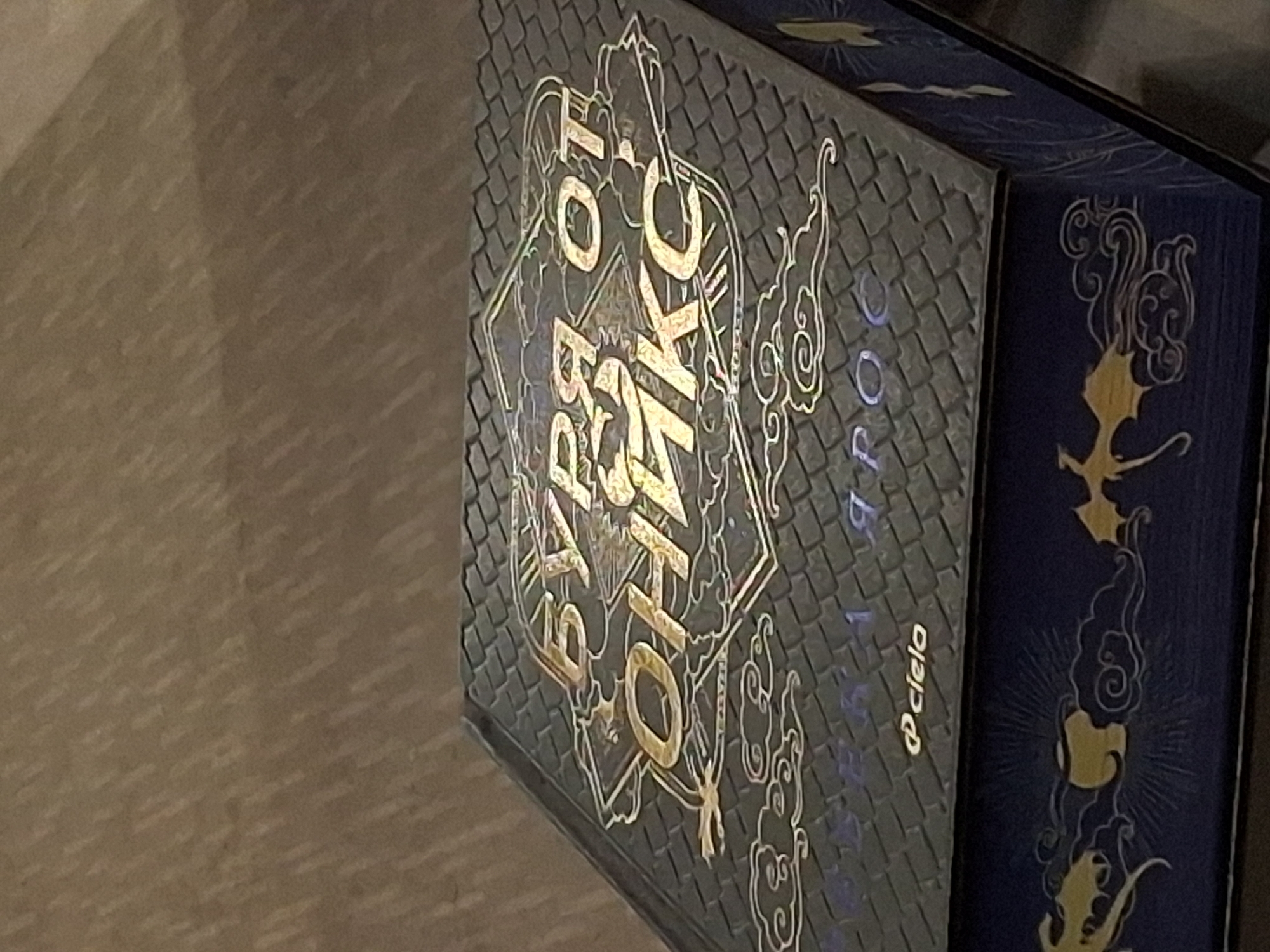 A dark blue or black rectangular object, likely a collector's edition book or a box containing a book, is centrally positioned and angled slightly from bottom left to top right. Its top surface features a textured, possibly scaled or tiled pattern, with prominent gold-colored Cyrillic text reading "БУРЯ ОТ ОНИКС" (Storm of Onyx) in an ornate, framed design. Below this, smaller gold Cyrillic text indicates the author "ДЖЕНИФЕР Л. АРМЕНТРАУТ" (Jennifer L. Armentrout) and "БУК 1. ЯРОСТ" (Book 1. Rage), followed by the publisher's logo "@ciela". The visible side of the object is deep blue and adorned with gold illustrations of stylized dragons or mythical creatures amidst swirling cloud motifs, centered around a radiant, sun-like or moon-like emblem. The object rests on a light brown, possibly wooden or carpeted, surface exhibiting a diagonal grain or patterned texture. The background above the object is out of focus, maintaining the same light brown hue and texture. No individuals are present. The precise location within Varna, Bulgaria, cannot be determined from the visual information provided in the image.