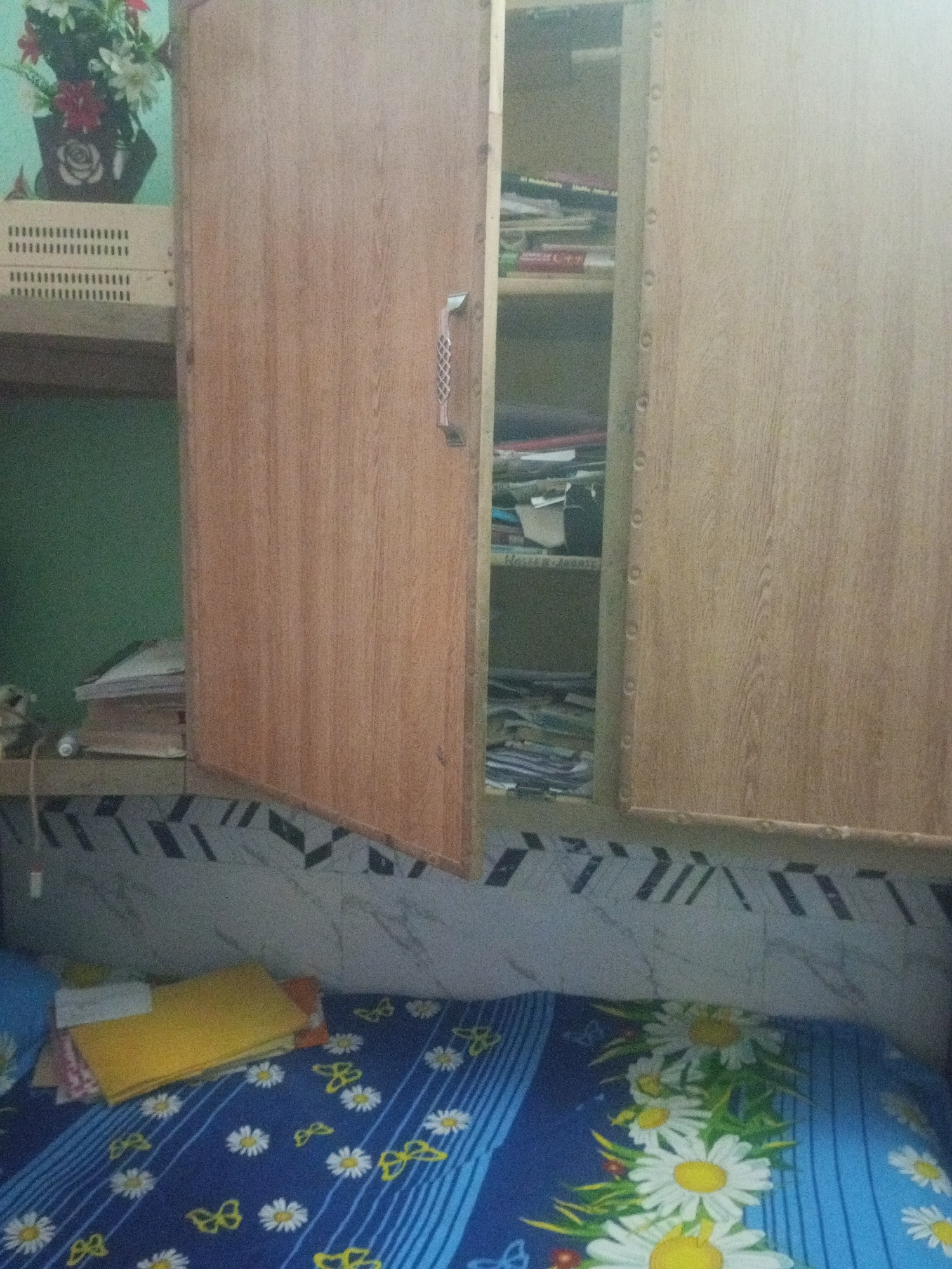 The image depicts an indoor scene, likely a bedroom, with a focus on a wooden cabinet. The cabinet is partially open, revealing shelves filled with books and papers. The exterior of the cabinet is made of wood-grained laminate. To the left of the cabinet, a shelf holds a floral arrangement in a dark vase and two white plastic containers. Below the cabinet, a bed is visible, covered with a blue bedsheet adorned with white daisies and yellow butterflies. The wall behind the bed has a patterned tile design. The overall impression is that of a lived-in domestic space. The image's content does not provide any direct clues to its specific location in Mansehra, Pakistan, or any details about people, actions, or interactions.