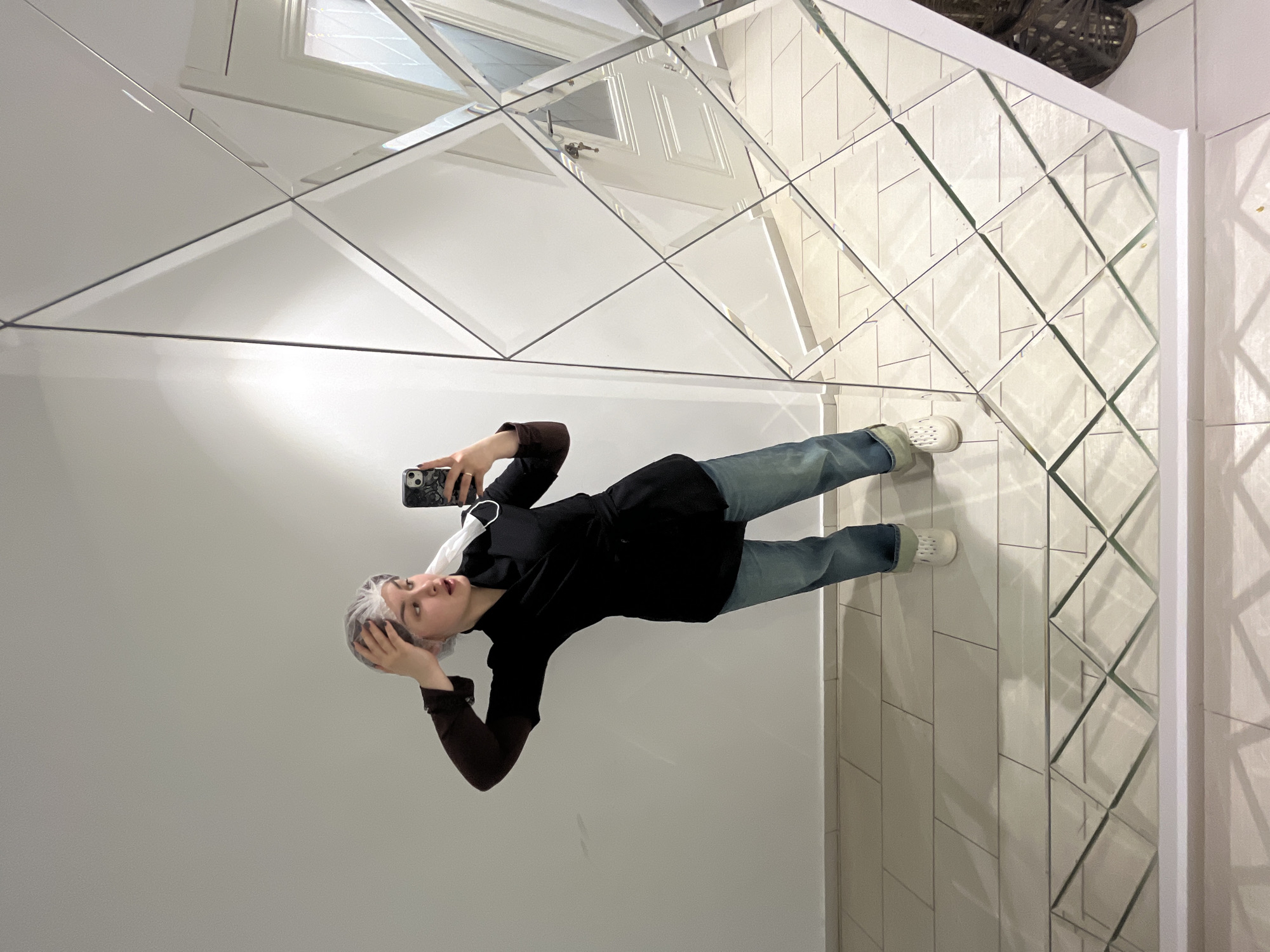 The image captures a young female taking a mirror selfie indoors in Neftekamsk, Russia. She is positioned slightly left of center, leaning forward with her left hand touching a clear disposable hairnet on her head. A white disposable face mask hangs from her right wrist. She wears a dark brown long-sleeved shirt under a black wrap-style tunic, light blue cuffed jeans, and white perforated clogs. Her right hand holds a black smartphone with a patterned case, pointed towards the mirror. The immediate environment includes a plain white wall and light-colored square tiled floor. A large, floor-to-ceiling mirror, framed in white and composed of numerous triangular and quadrilateral reflective segments, occupies the right side of the scene. Reflected in the mirror on the far right is a white door with a window featuring a diamond-patterned pane and a visible metallic door handle with a key. A dark, woven object is partially visible on the floor to the far right. The area is illuminated by bright interior lighting.