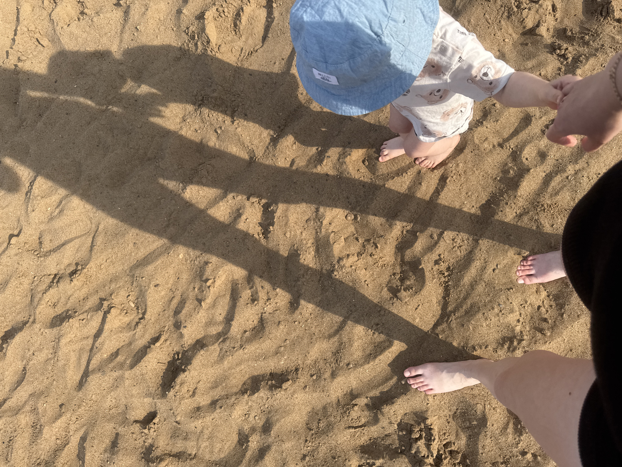 The image captures a close-up, overhead view of sandy ground with distinct shadows suggesting the presence of at least two people. On the left, a bare foot and lower leg are visible, indicating an adult. To the right, a child is partially visible, wearing a light blue hat and shorts adorned with a bear pattern. The child's feet are also bare. The hands of two individuals, presumably the adult and child, are clasped together, signifying an interaction. The environment appears to be a sandy area, possibly a beach or a sandpit, with ripples and textures in the sand indicating it may have been recently disturbed or shaped by wind or water. The lighting suggests it is daytime, with the sun casting long shadows across the scene. The location is Chaikovsky, Russia, though specific landmarks are not present in the image.