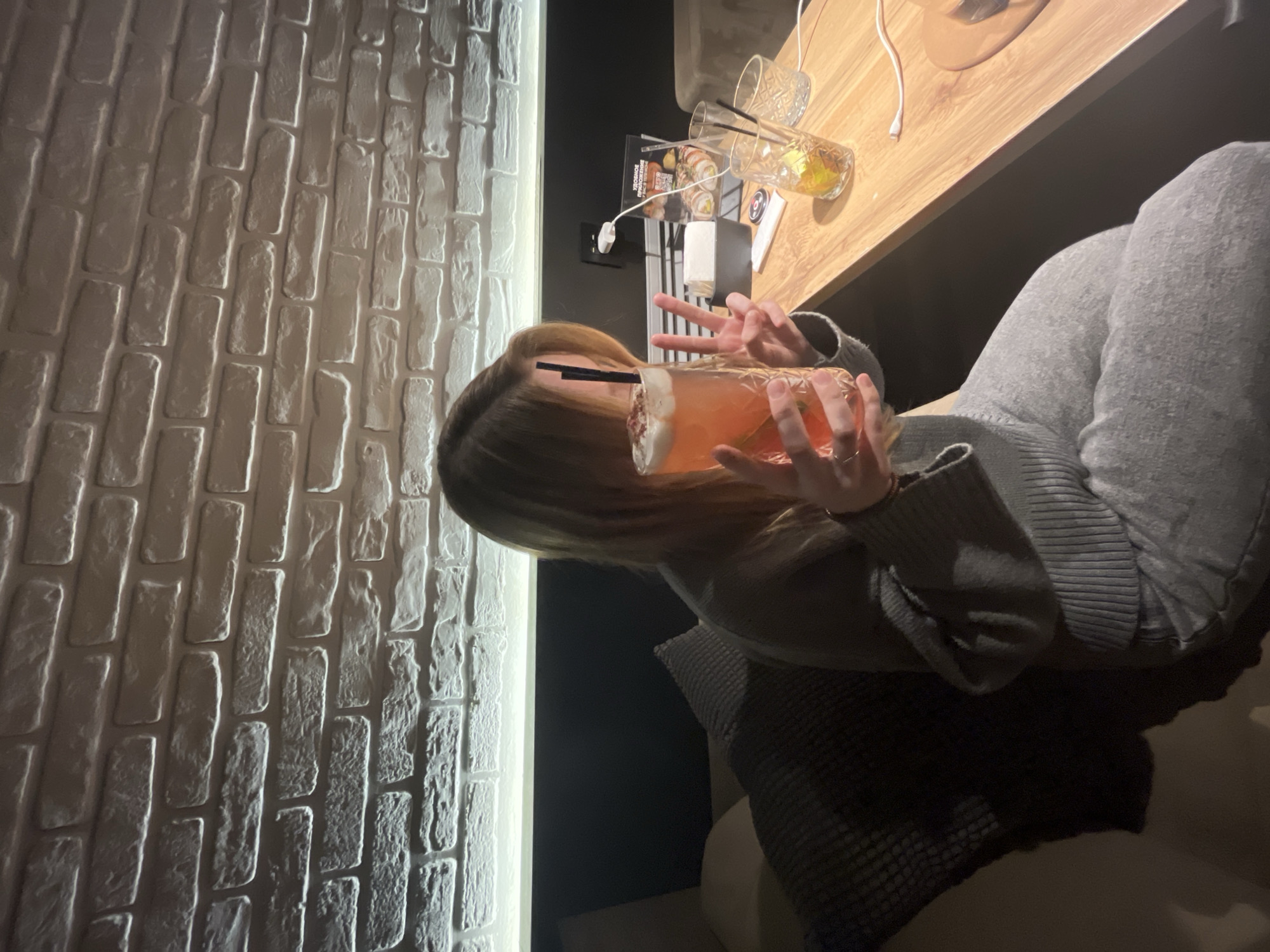 A person is holding a cocktail glass with both hands, making a peace sign with their left hand. They are seated, with their knees bent and covered by gray pants. The room has a brick wall painted white on the left. A table is on the right with glasses and a menu. The city is Chaikovsky, Russia.