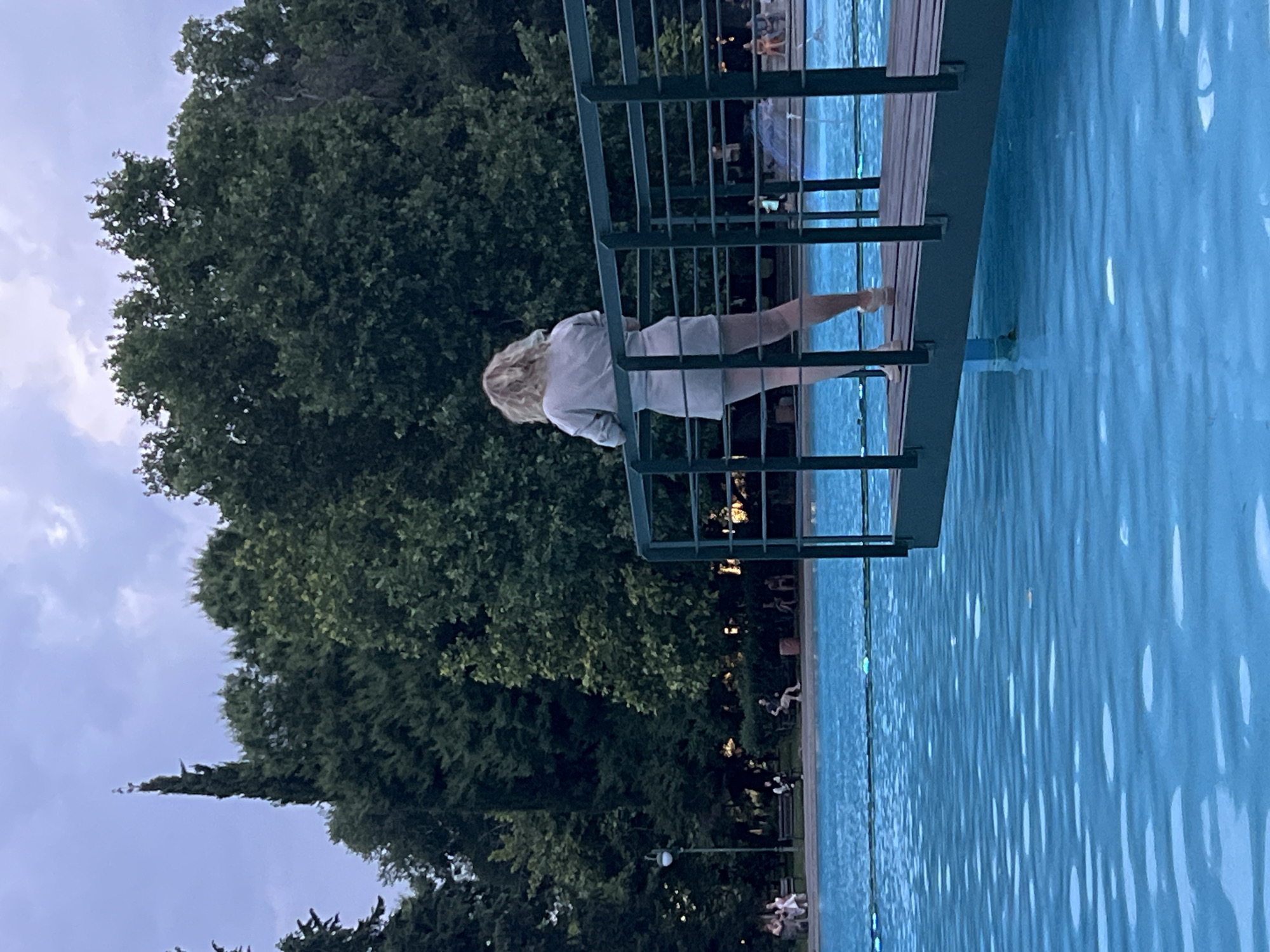 A woman with blonde hair, viewed from behind, stands on a wooden walkway with a dark green railing that extends over a bright blue body of water. She is wearing a light-colored short dress and white shoes. The walkway is positioned in the foreground, with the water filling the lower half of the image. In the background, a dense line of dark green trees borders the water. Further back, several people are seated on benches under the trees, and a lamppost is visible on the left side. The sky above is overcast with gray clouds. The scene appears to be within the Sea Garden in Plovdiv, Bulgaria, specifically near the Neptune fountain.