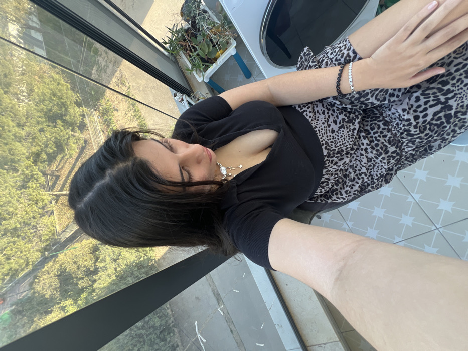 The image displays a female subject captured from an elevated, overhead perspective, seated or leaning indoors in Kiryat Bialik, Israel. She possesses long, dark hair and a light complexion, gazing downwards. Her right arm is extended forward, suggesting a selfie pose. She wears a black short-sleeved collared top with a deep V-neck and a light-toned leopard print skirt. Adornments include a necklace with a central pearl/oval stone, small stars, and a cross, along with a black beaded bracelet and a silver-colored rectangular link bracelet on her left wrist. Behind her, large vertical windows offer a view of an outdoor environment consisting of green trees and foliage, with patches of visible ground. The indoor area features a light-colored tiled floor with a distinct geometric pattern. To the right of the subject, a white planter containing various green succulent plants rests on a blue stand near the window, adjacent to a white surface or object. The scene is illuminated by natural sunlight.