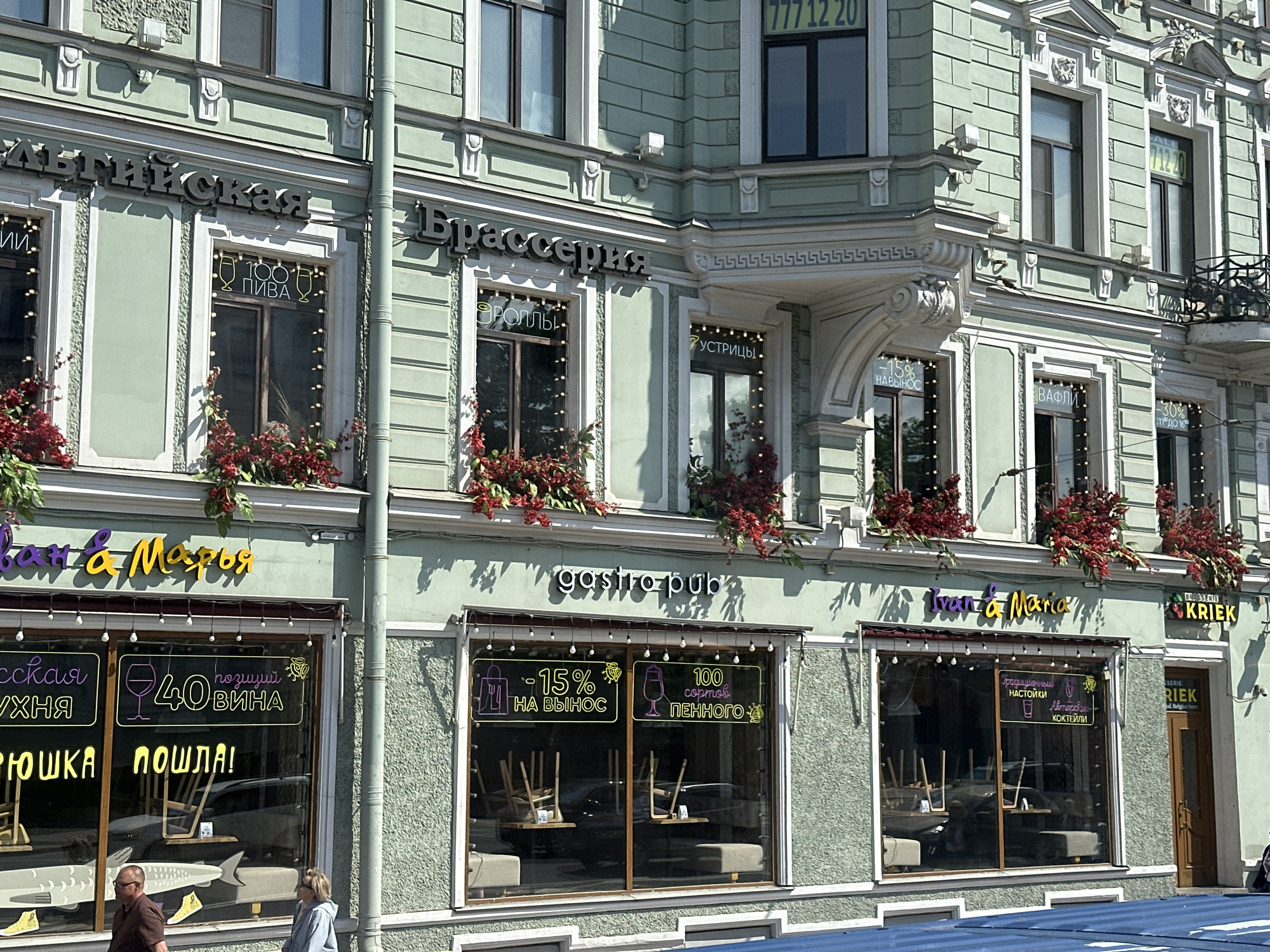 The image shows a building facade in Saint Petersburg, Russia, with various businesses on the ground floor. The architecture is classical, with decorative molding and a light green color scheme. The businesses include a restaurant called "Baltiyskaya Brasserie" (Балтийская Брассерия), and establishments named "Ivan & Maria" and "gastro-pub". Each business features large windows displaying neon signs and menu items. Floral arrangements are visible above the windows. Pedestrians walk past the storefronts. The image is taken during the day.