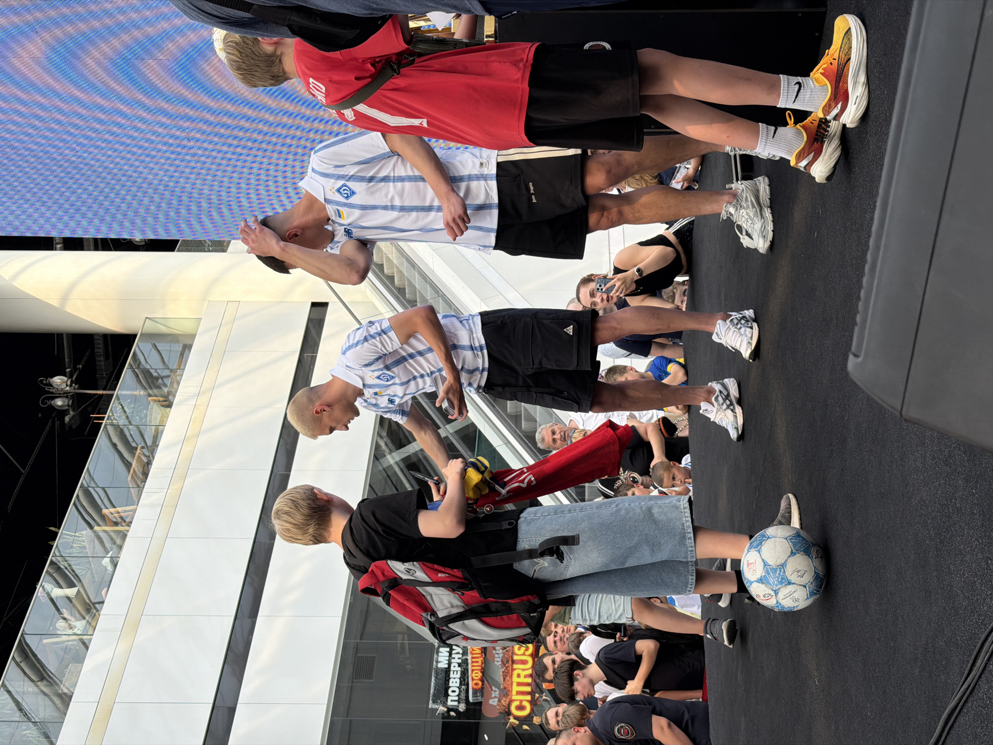 In Kyiv, Ukraine, on an outdoor stage in front of a large LED screen, a group of people are gathered. In the foreground, a young person wearing a black t-shirt and denim shorts stands with a red and gray backpack. Beside them is a soccer ball with blue and white panels. To the right, two young men in white and blue striped athletic jerseys and black shorts are interacting. One of them is reaching out to the person with the backpack, while the other is touching his head. The background shows a modern building with multiple levels and a large blue LED screen displaying abstract patterns. A crowd of onlookers, including adults and children, are visible behind the main subjects, some holding up phones to take pictures. The atmosphere appears to be a public event or gathering.