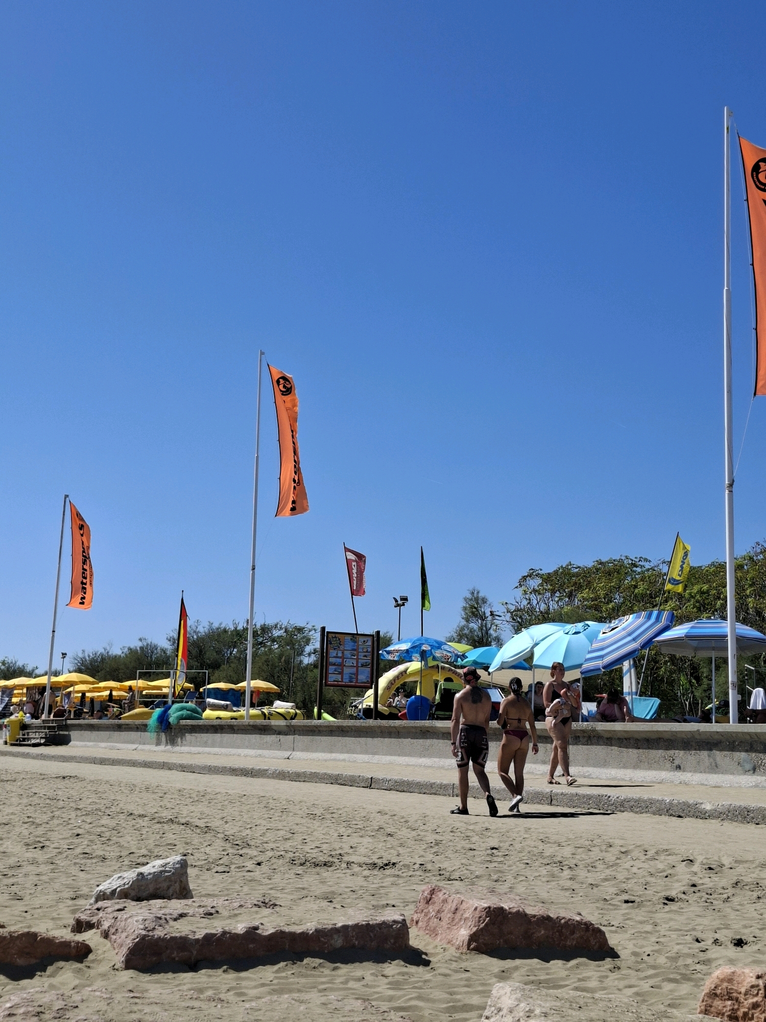 The image depicts a sunny day on a sandy beach in Caorle, Italy. A paved walkway runs parallel to the beach, separating it from a row of beach umbrellas and trees. Several orange flags with black lettering are prominently displayed on tall poles, indicating "water sports" and a company logo. A few individuals are visible walking on the paved path. In the foreground, large rocks are scattered on the sand. The sky is clear and blue.