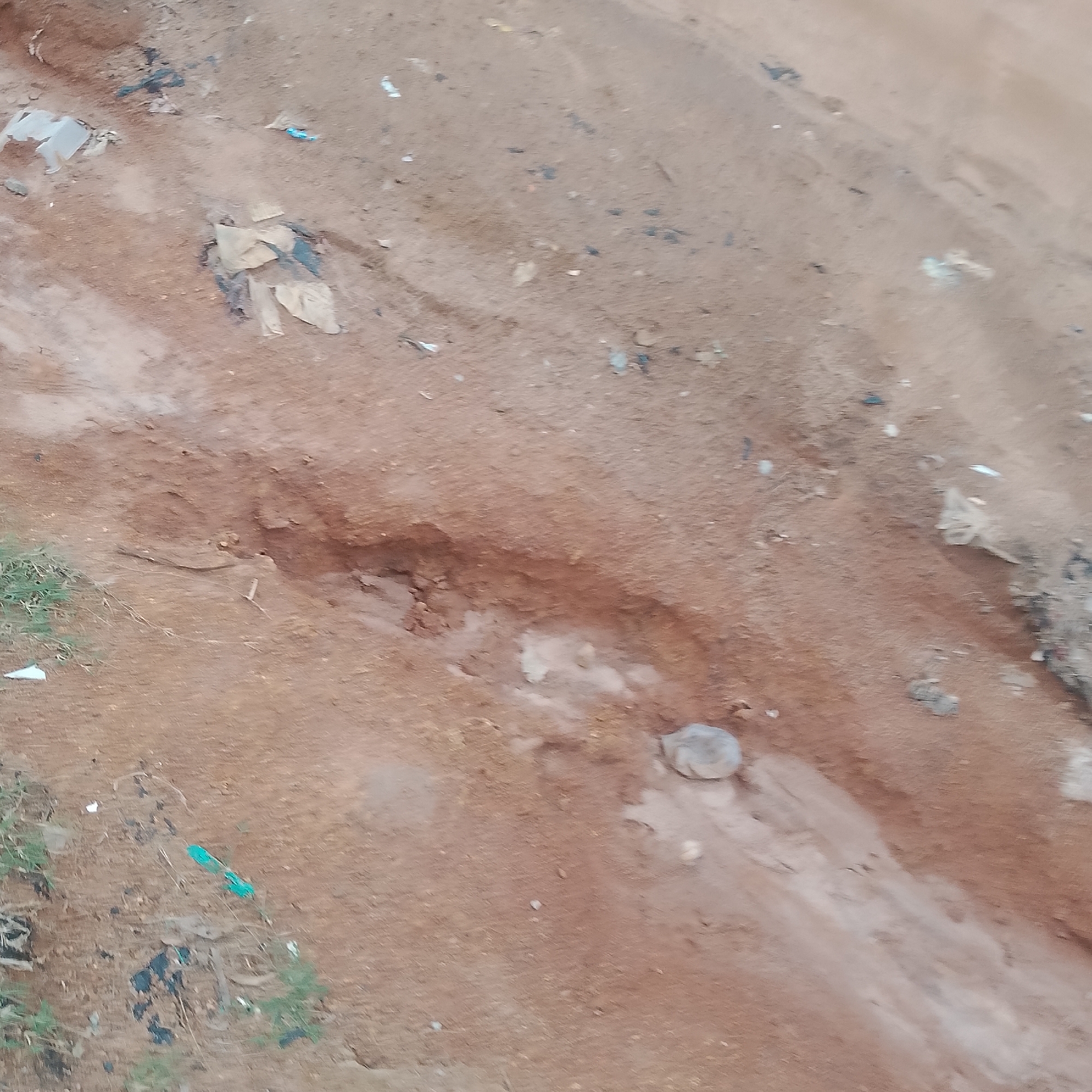 The image depicts a dirt path or road in Jalingo, Nigeria. The ground is composed of reddish-brown soil with some lighter patches, suggesting the presence of water or a different soil composition in certain areas. There is a noticeable erosion gully cutting across the path, indicating potential drainage issues or heavy rainfall. Scattered across the surface are various pieces of debris, including what appear to be plastic bags, torn fabric, and other unidentifiable waste. Small patches of green grass and dry vegetation are visible along the edges of the path. The scene appears to be an outdoor, unpaved area within the city, possibly an informal road or track. There are no people visible in the image.