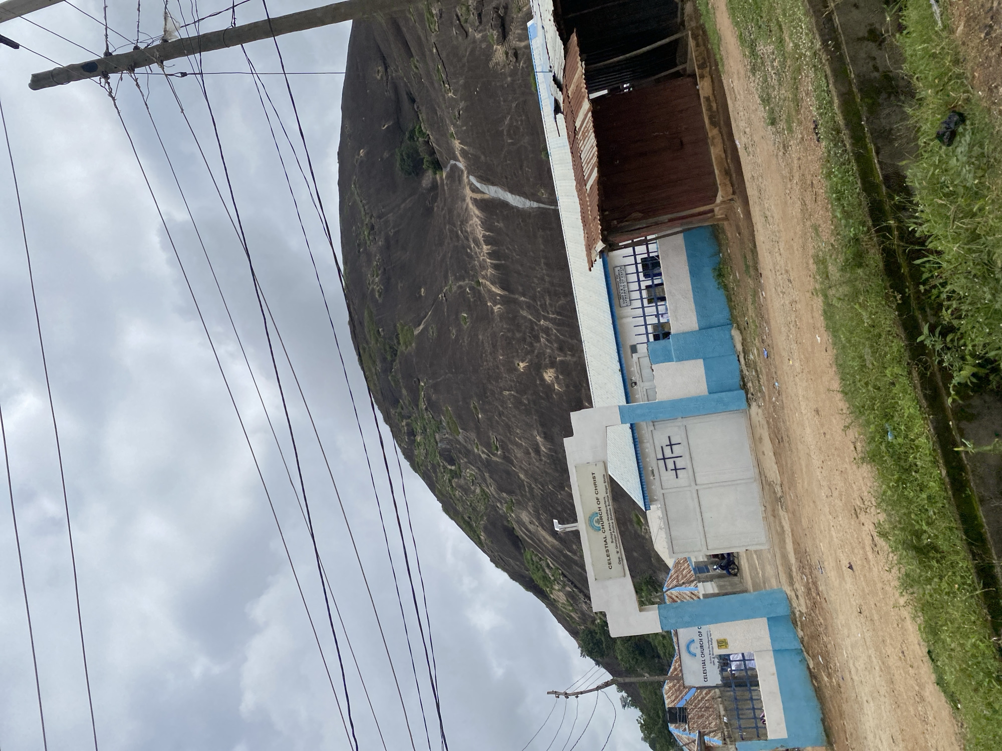 The image depicts the Celestial Church of Christ in Suleja, Nigeria, situated at the base of a large, rocky hill with vegetation. In the foreground, a dirt road leads to the church's entrance, marked by a white archway with blue trim. Above the archway, a sign reads "CELESTIAL CHURCH OF CHRIST" with smaller text below indicating "Suleja Area Headquarters, Opp. B' Division Police Station, Suleja, Niger State." The church building is painted white with blue accents, featuring a large white gate with a cross symbol. To the right of the church entrance, a small, dilapidated structure made of corrugated metal stands. Overhead, electrical wires crisscross the sky, supported by a concrete pole on the right side of the frame. The sky is overcast with gray clouds. A few people are visible entering the church through the main gate, and a motorcycle is parked inside the premises. The overall environment appears to be a mix of established religious infrastructure and informal local structures, set against a natural landscape dominated by the imposing hill.