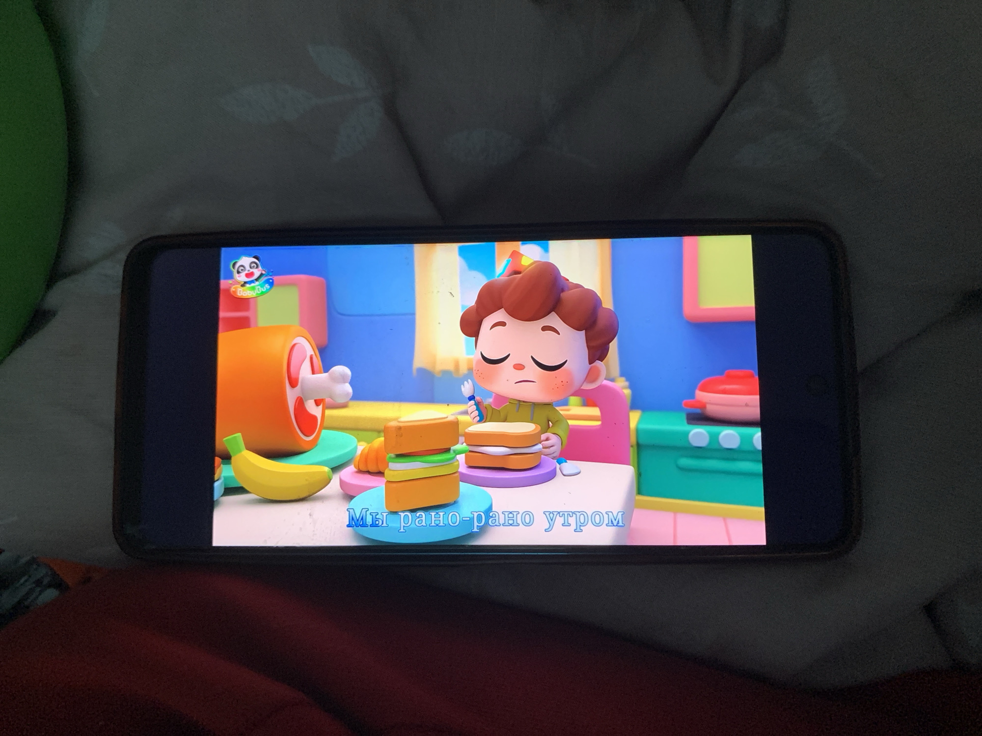 The image shows a smartphone displaying a cartoon. The cartoon features a character with red curly hair and big eyes, holding a fork, and a sandwich. There is also a banana, a ham bone, and a baby panda with a colorful halo. The words "Мы рано-рано утром" are visible on the screen, which translates to "We are early in the morning." The smartphone is placed on a patterned surface. The image is taken from a slightly overhead angle, with the smartphone rotated sideways. The scene appears to be an indoor environment, likely a home.