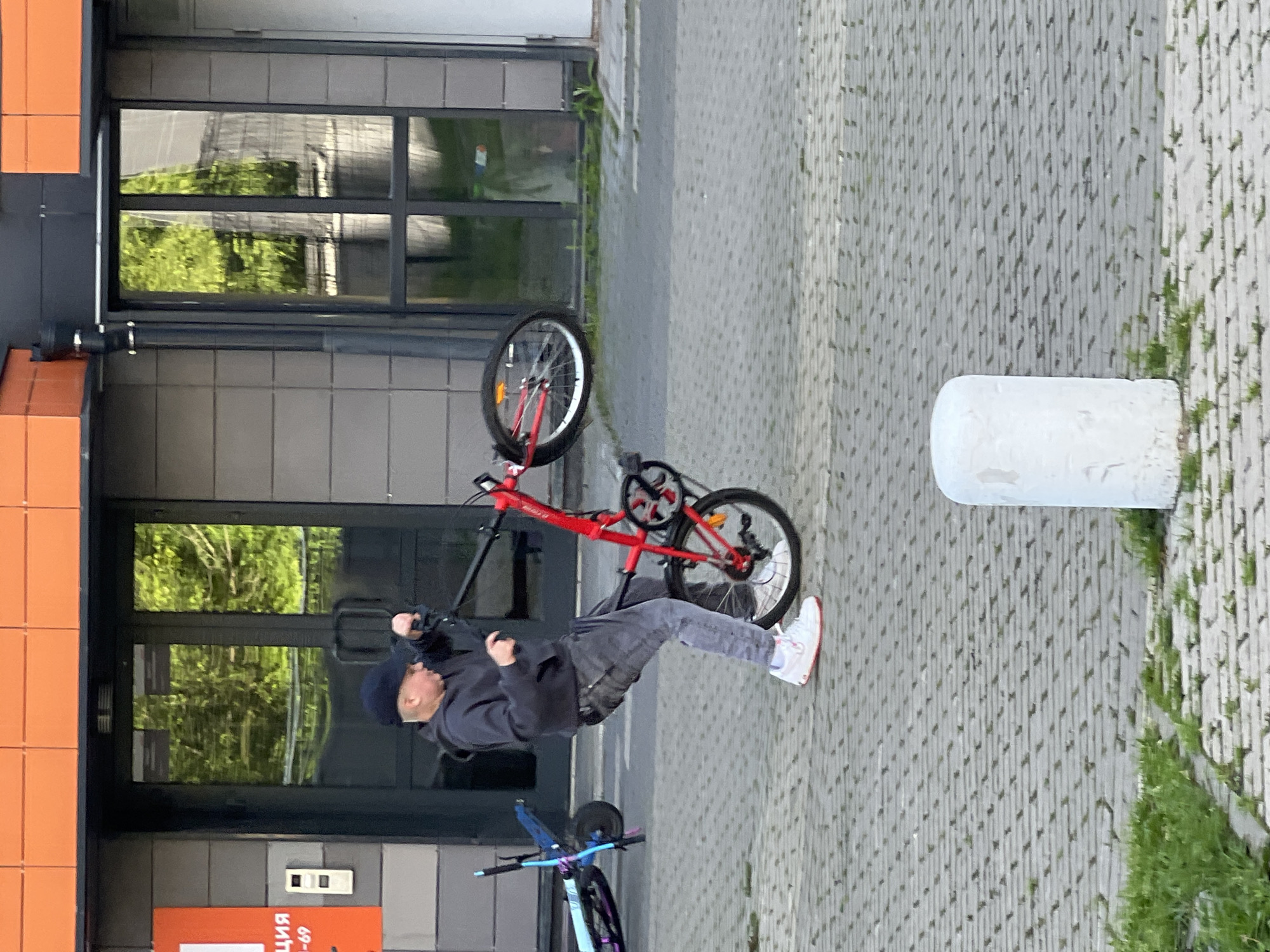 A person is performing a bicycle trick, balancing on the rear wheel of a red bicycle while the front wheel is elevated. The individual is wearing a dark hooded sweatshirt, grey pants, and white sneakers with red accents. They have a dark blue beanie on their head. The bicycle has a black seat and handlebars, with silver rims and black tires.
The scene takes place on a paved area with a grey, patterned surface, possibly a sidewalk or plaza. To the left, there is a building with large windows reflecting green foliage, suggesting trees or other vegetation. The building has sections with orange and grey tiling. A white cylindrical bollard stands to the right of the frame. In the background, another bicycle, primarily blue and black, is visible, resting upright.
The lighting suggests it is daytime, possibly overcast, as there are no harsh shadows. The overall mood appears to be one of action and skill, focused on the bicycle stunt.
Visible text: On an orange sign in the lower left portion of the image, Cyrillic text is partially visible, reading "ЦИЯ" and "69-69". This suggests the location is in Russia, as indicated by the Cyrillic alphabet.
