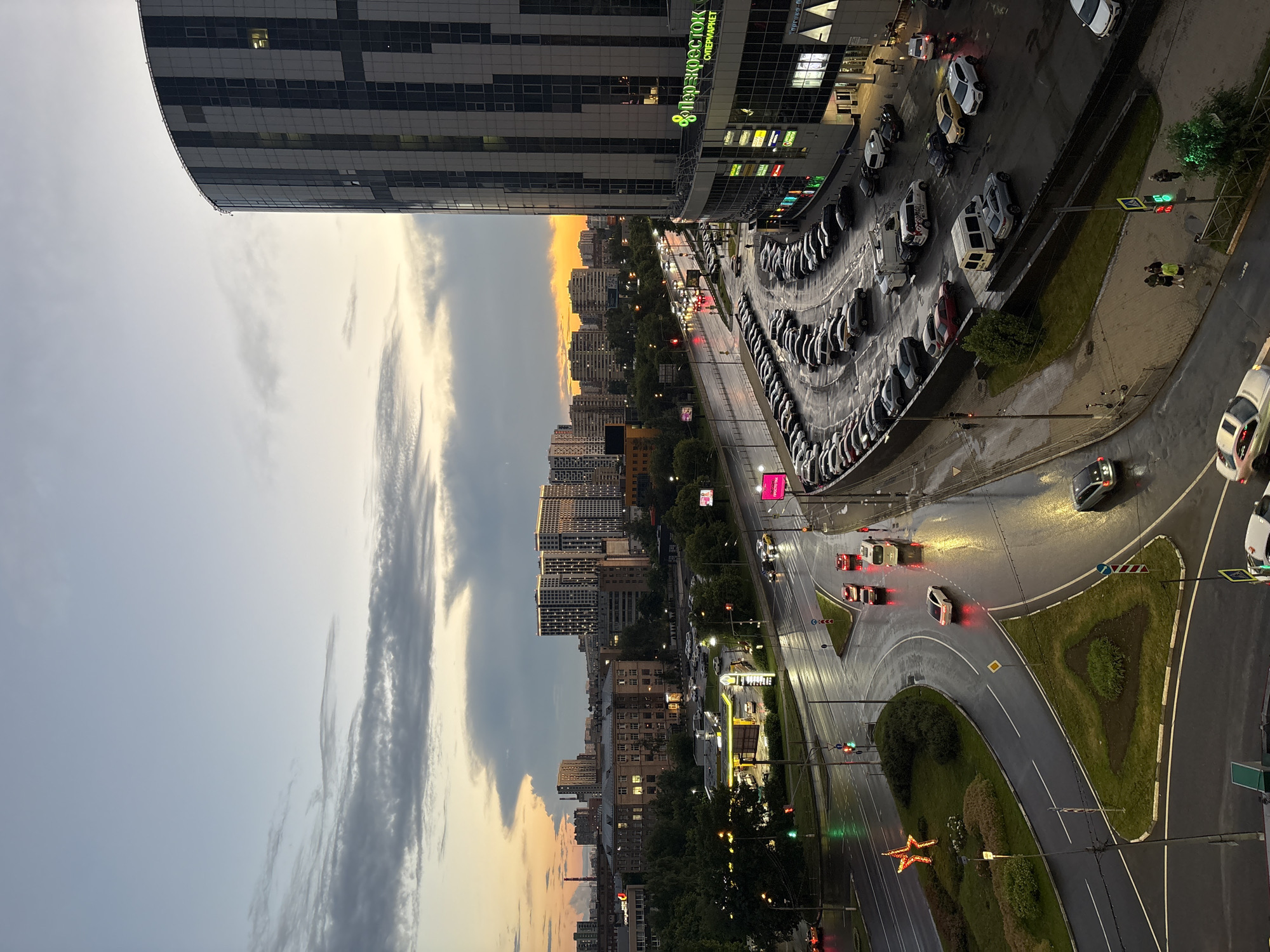 This is an aerial photograph of a street intersection and a parking lot at dusk in the city of Lesnye Polyany, Russia. The sky exhibits a gradient of colors from pale blue at the top to orange and gray clouds near the horizon, suggesting the sun has recently set. A large, modern, curved building with a dark facade and vertical white and gray panels dominates the right side of the frame. The building has a sign in Russian that reads "ПЕРЕКРЕСТОК СУПЕРМАРКЕТ".
The street is wet, indicating recent rain, and reflections of lights from buildings and vehicles are visible on the asphalt. Several cars are on the road, some appearing to be moving, while others are parked. Traffic lights are visible and illuminated, with one showing green.
A large, multi-row parking lot is situated in front of the curved building, filled with numerous cars of various colors and models, mostly parked. A smaller road with a star-shaped decorative light fixture on a green median is to the left, curving away from the main intersection. In the background, multiple apartment buildings and other structures are visible against the darkening sky, with some windows illuminated.
A few individuals are visible on the sidewalks and near the parking lot, appearing to be walking or standing. The overall scene depicts a commercial and residential urban environment during twilight, with active traffic and a busy parking area.