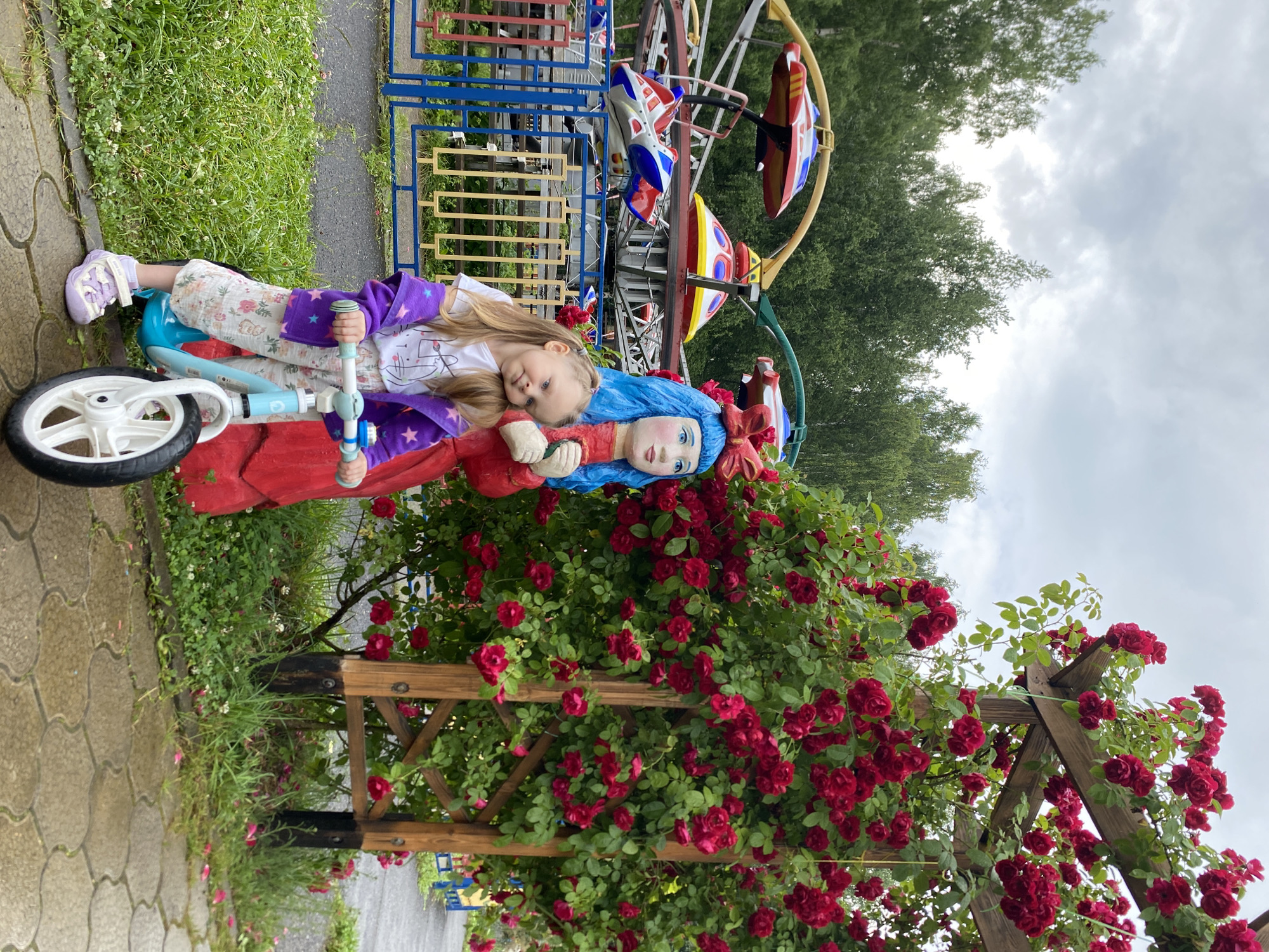 A young girl with blonde hair is posing for a picture on a balance bike in front of a large bush of red roses. She is wearing a white t-shirt with a graphic print, a purple jacket with star patterns, white floral-patterned pants, and light purple sandals. Next to her stands a life-sized statue of a doll with blue hair, wearing a red dress and holding her hands in front of her. The balance bike is light blue and white. In the background, a playground carousel with colorful ride vehicles is visible, along with green trees and a cloudy sky. The ground is paved with stone tiles and grass. The scene appears to be an outdoor amusement area or park in Chaikovsky, Russia.