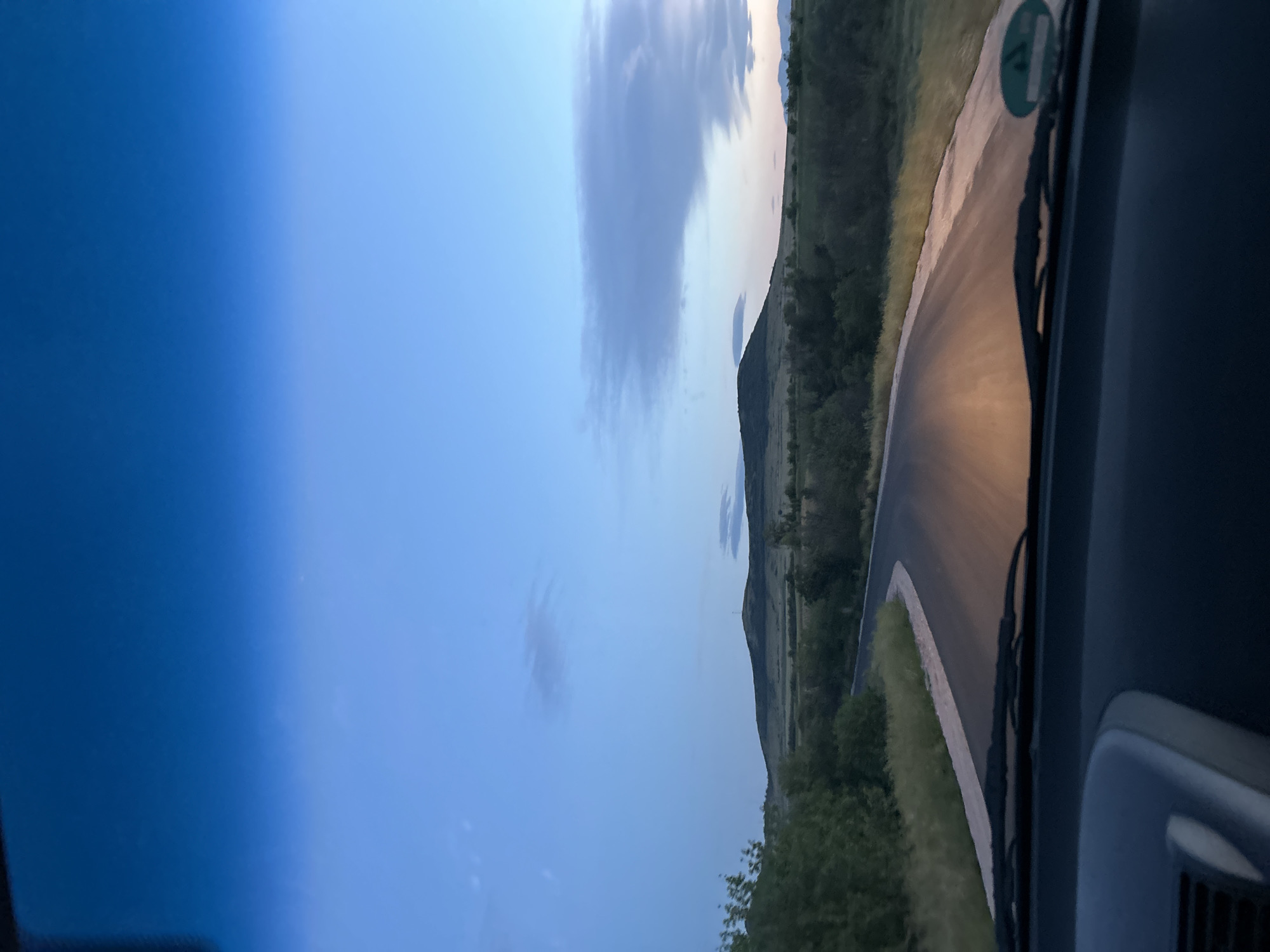 The image shows a view from inside a vehicle driving on a road outside of Ovchepoltsi, Bulgaria. The road curves to the right, flanked by green vegetation. In the distance, there is a mountain. The sky is a gradient of blue, with scattered clouds. The vehicle's windshield and dashboard are partially visible.
