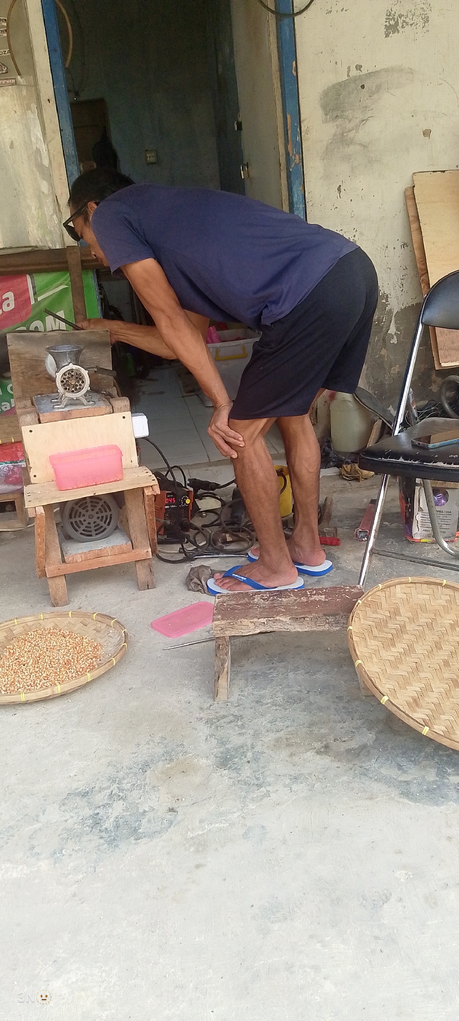 A person, seen from the side and slightly from behind, is bent over a meat grinder. The person is wearing a navy blue t-shirt and black shorts, with dark-rimmed sunglasses. Their hair is dark and shoulder-length. They are standing on a concrete floor in what appears to be an outdoor or semi-outdoor space, possibly a shop or workshop in Sukanegara, Indonesia. To the left of the meat grinder is a woven basket filled with corn kernels. The meat grinder is mounted on a wooden structure, and below that is an electric motor with a fan. In front of the person, on the floor, are a pair of blue flip-flops. To the right, a metal chair with a black seat and back is visible, and a woven tray rests on the ground. The background shows a wall with peeling paint and an open doorway revealing a dark interior. A yellow container or bottle is also present in the background. Electrical wires and a device with a digital display are connected to the motor.