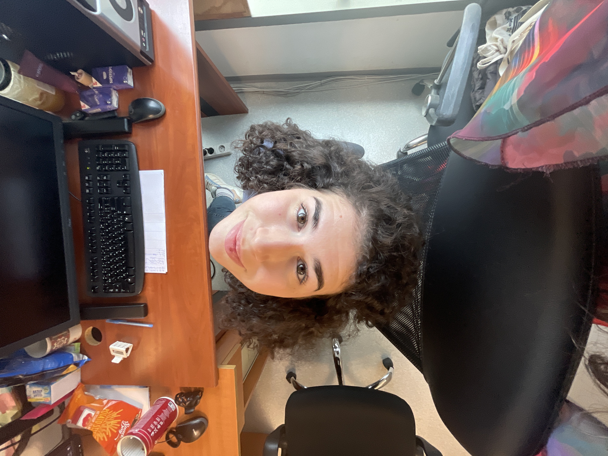 The image, taken from a low, upward-looking perspective and rotated, features a young woman with curly dark brown hair, light brown eyes, and fair skin. She is looking directly at the camera with a gentle smile, positioned to the right of a cluttered wooden desk. She is seated in a black office chair with a mesh back and silver armrests. A vibrant, multi-colored fabric, possibly a scarf or part of an garment, with patterns in red, purple, blue, and green, is draped over the chair's back. The woman is wearing blue jeans and light-colored sneakers with blue stripes.
On the wooden desk to her left, a black computer monitor and a black keyboard are prominent. A white piece of paper is visible under the keyboard, and a black computer mouse rests nearby. Above the desk, on a shelf, are silver and black speakers or a computer tower, and two small purple boxes labeled "SUPERSOFT," likely tissue boxes. The desk also holds a red Pringles can, an orange packet with "POCKY" visible, a white paper cup, a black pen, and a small white stapler. A circular hole in the desk suggests cable management. The background shows a light-colored wall with visible electrical wires and standard European power outlets, placing the scene indoors, likely in an office or study room in Sofia, Bulgaria. The shot appears to be a casual portrait or selfie in a personal workspace.