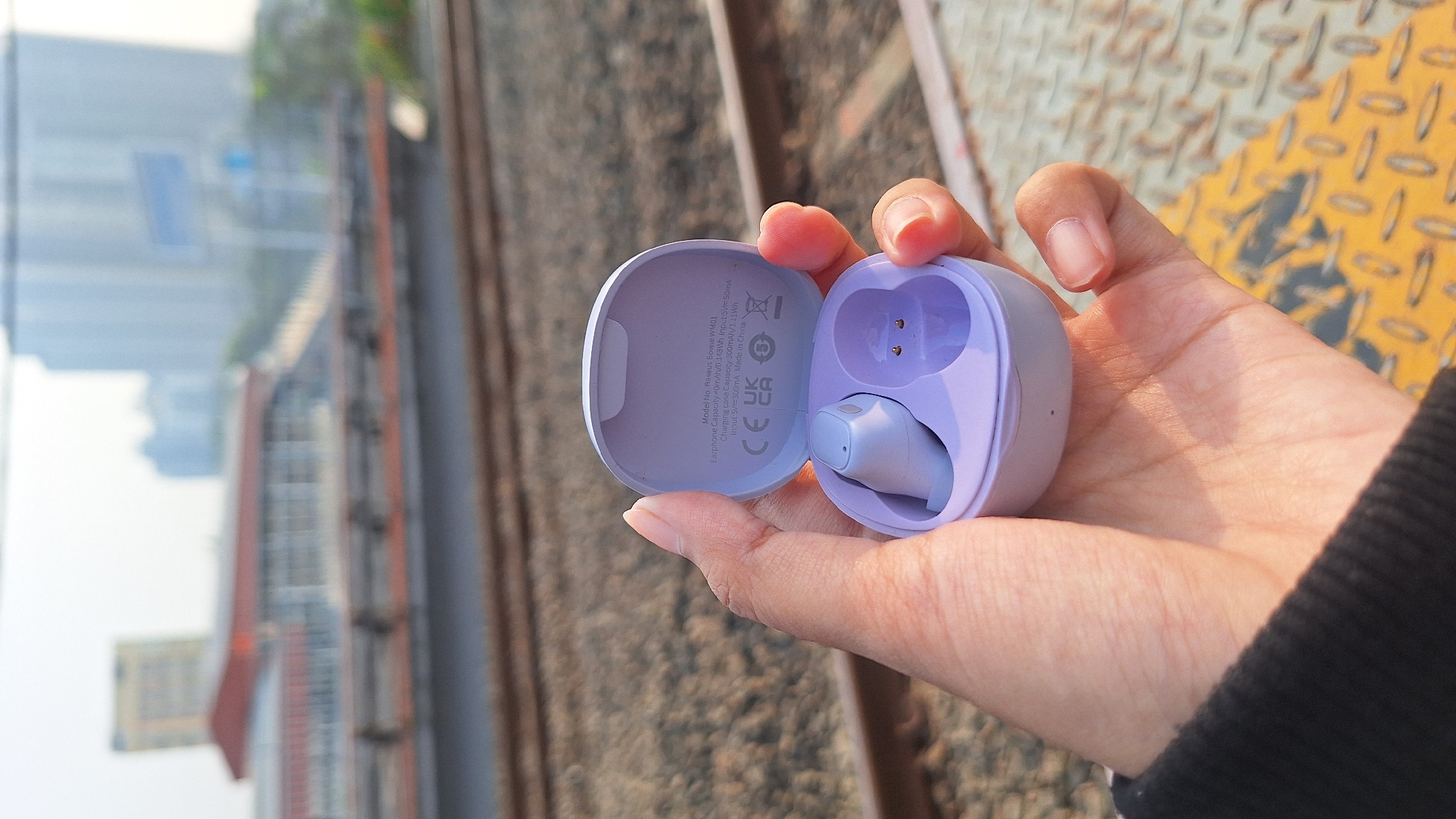 In North Jakarta, Indonesia, a hand holds an open, light purple wireless earbud charging case. Inside the case, one light purple earbud is visible. The charging case is positioned in front of a blurred background depicting train tracks and distant city buildings. The person's hand, partially visible, is wearing a black garment. The overall lighting suggests daylight.