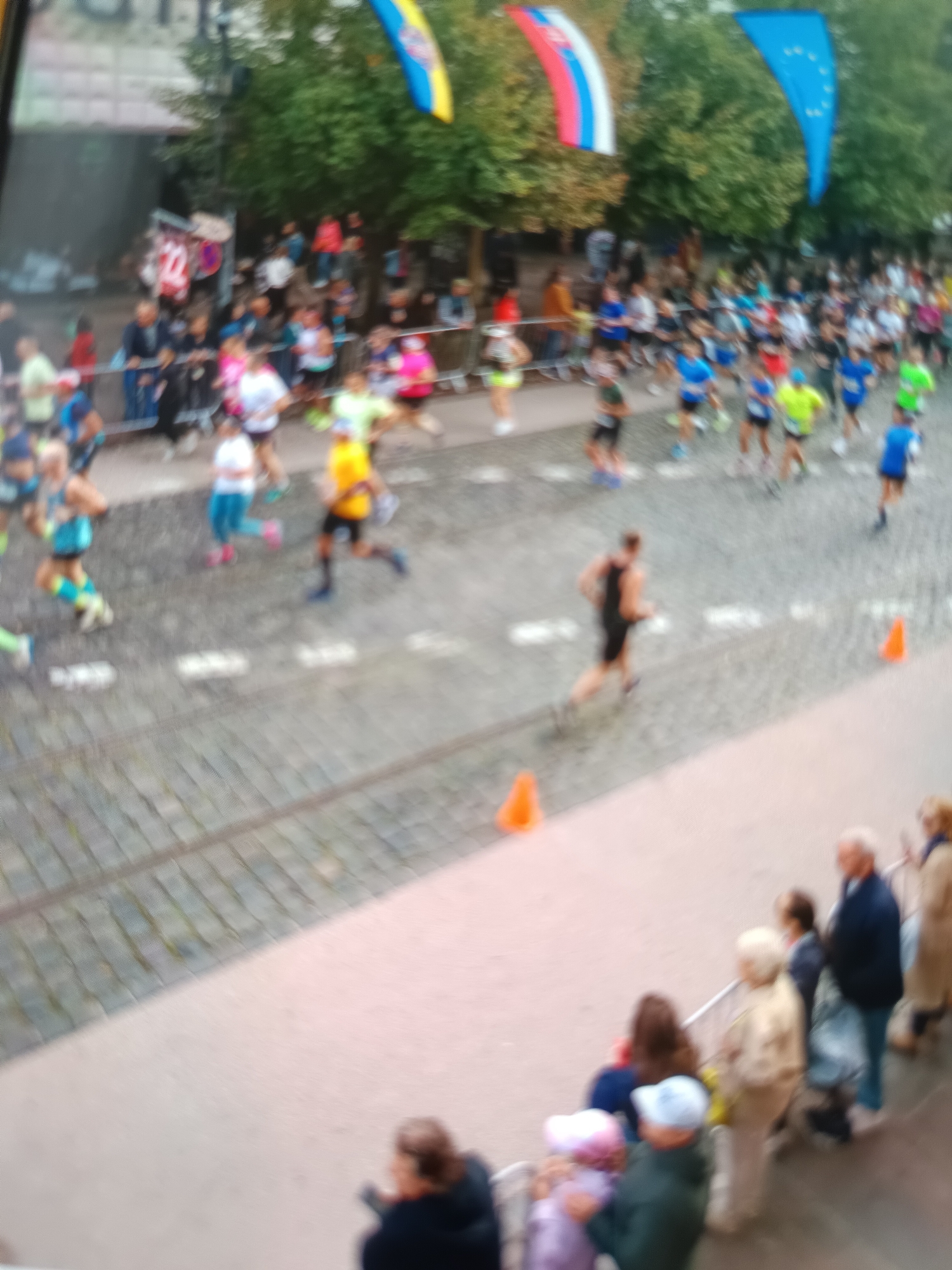 This media file depicts an outdoor running race, likely a marathon or a similar event, taking place on a cobblestone street. The image is blurry, suggesting movement and a candid shot. The foreground shows a crowd of spectators lining a barrier, observing the runners. People of various ages are visible, some in casual attire, others in athletic wear.
The main subjects are the runners in motion, some wearing brightly colored athletic outfits, while others are in darker clothing. They are spread across the street, indicating a significant number of participants. Orange traffic cones are placed along the course, marking the path.
In the background, there are trees and buildings, suggesting an urban setting. Flags are visible, including what appears to be the Slovak flag and a blue flag with stars, possibly the European Union flag, hinting at the location in Košice, Slovakia. The overall lighting suggests it might be daytime, possibly overcast given the lack of strong shadows. The mood appears to be energetic and communal, with people gathered to participate in or watch the event. There are no clear, readable text elements in the image.