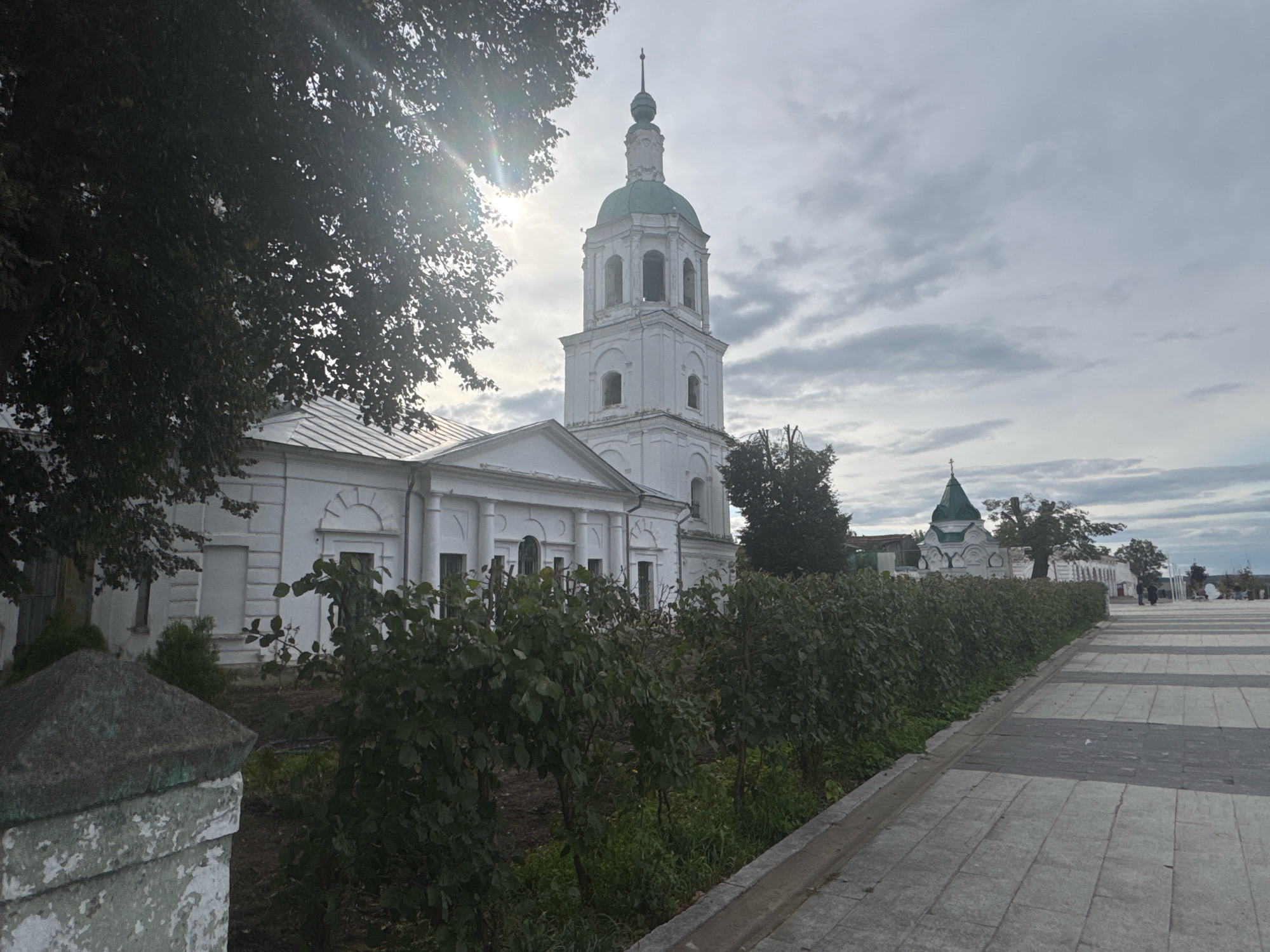 The image depicts the John the Baptist Monastery in Zaraysk, Russia, under an overcast sky. In the foreground to the left, a weathered stone pillar stands partially obscured by overgrown green bushes. Adjacent to the bushes, a paved walkway with a checkered pattern of light and dark grey tiles extends to the right. To the left of the walkway, a long, neatly trimmed hedge runs along the perimeter of a white building. This building features a symmetrical facade with multiple windows and decorative elements above the doorways. Dominating the background is a tall, white bell tower with a green onion dome, topped with a spire. To the right of the bell tower, another smaller white building with a green roof is visible, partially hidden by trees. Several people are sparsely scattered along the walkway in the distance, some appearing to be walking or standing. The overall scene suggests a quiet, historical location.