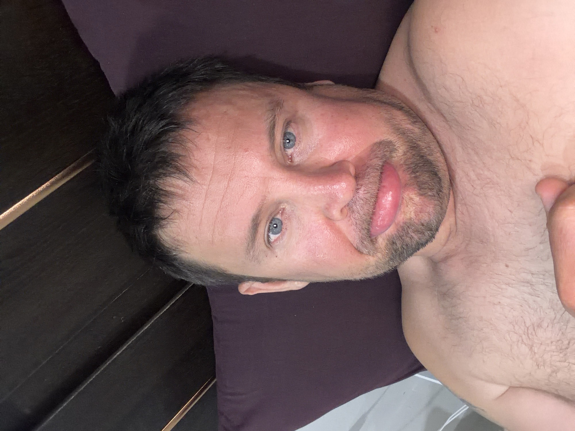 A close-up, eye-level, overhead shot shows a fair-skinned man with short, dark hair, blue eyes, and a beard. He is lying down, appearing to be in bed, with a dark brown headboard and a dark purple pillow visible in the background. The man is partially undressed, with his shoulders and chest exposed. His skin appears flushed, and his mouth is open with his tongue slightly protruding, giving a playful or suggestive expression. The lighting is direct, highlighting his features and the texture of his skin and hair. The scene is a personal, indoor setting, likely a bedroom, given the presence of a bed and pillow. There is no information about the city of Baan Mae Nam, Thailand in the image.