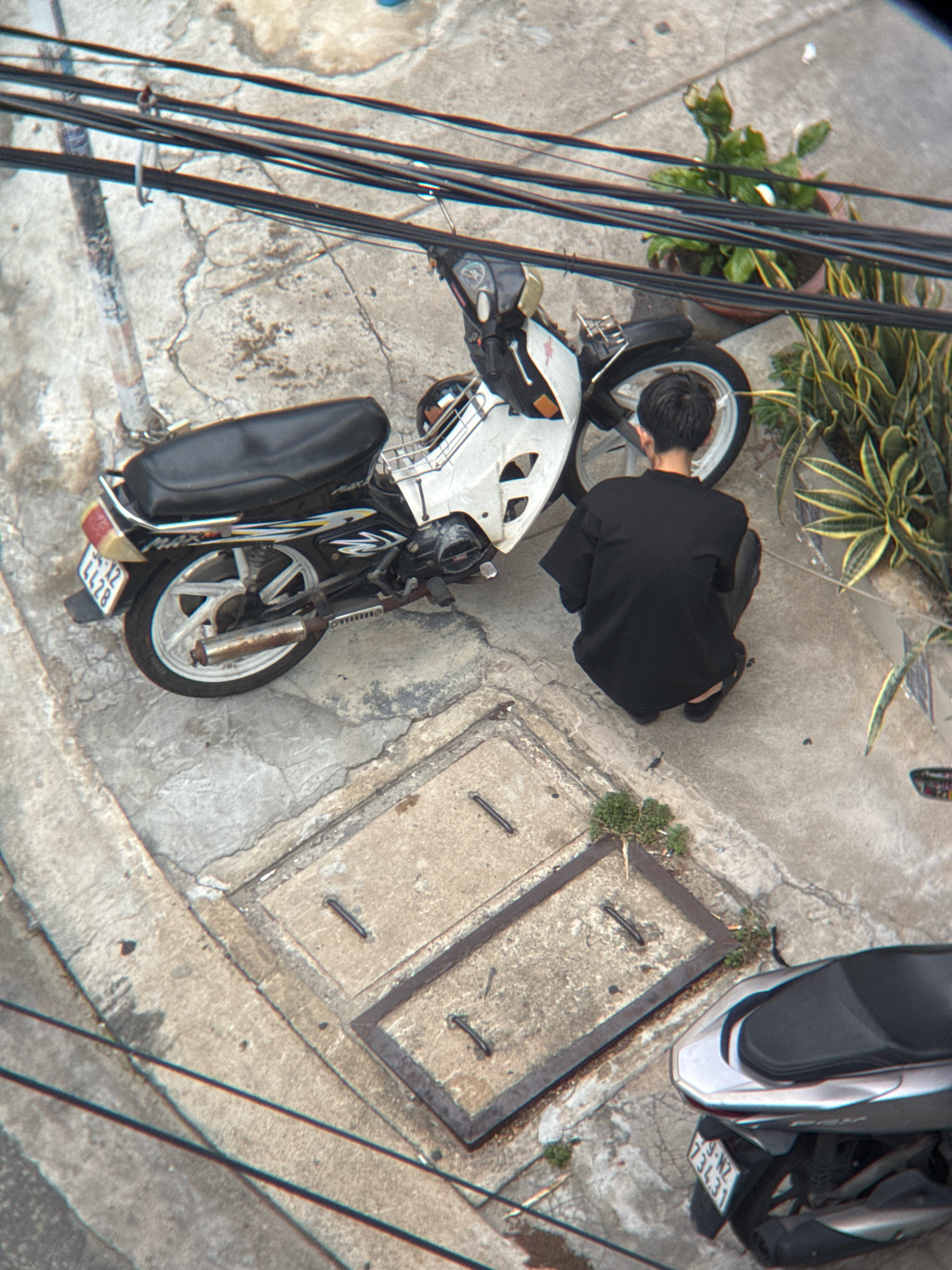 The image captures a street scene in Nha Trang, Vietnam, looking down at an angle. The ground is paved concrete, with cracks and a manhole cover visible. A white and black motorcycle is parked on the left, and a silver scooter is on the right. A young man wearing a black t-shirt and dark pants is squatting beside the white motorcycle, facing away from the camera. He appears to be looking at something on the ground or the motorcycle. Several electrical wires are strung overhead, crossing the frame. Green plants are growing in pots on the right side of the image, and a utility pole is visible on the left. The scene suggests an everyday moment in an urban environment.