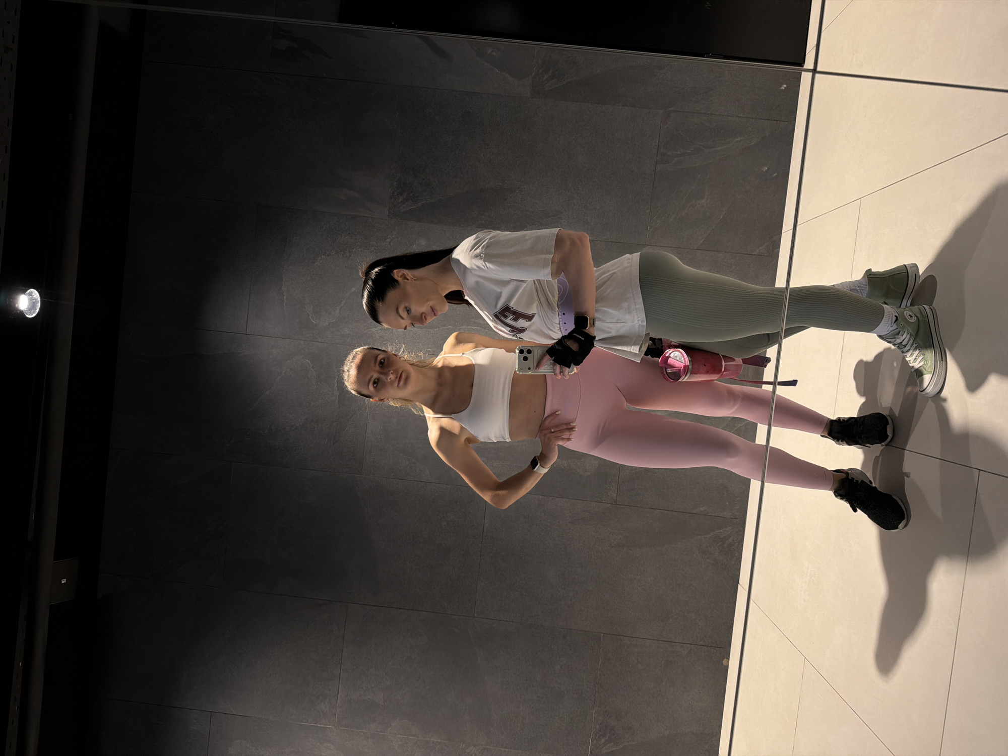 Two women in athletic wear stand in a room with dark grey textured walls. The floor is light-colored tile. The women are posing and taking a selfie. One woman is wearing a white sports bra and pink leggings. The other woman is wearing a white t-shirt and light green leggings. A pink water bottle and a cell phone are visible. The location is potentially a gym or fitness studio in Perm, Russia, indicated by the username on the phone’s screen.