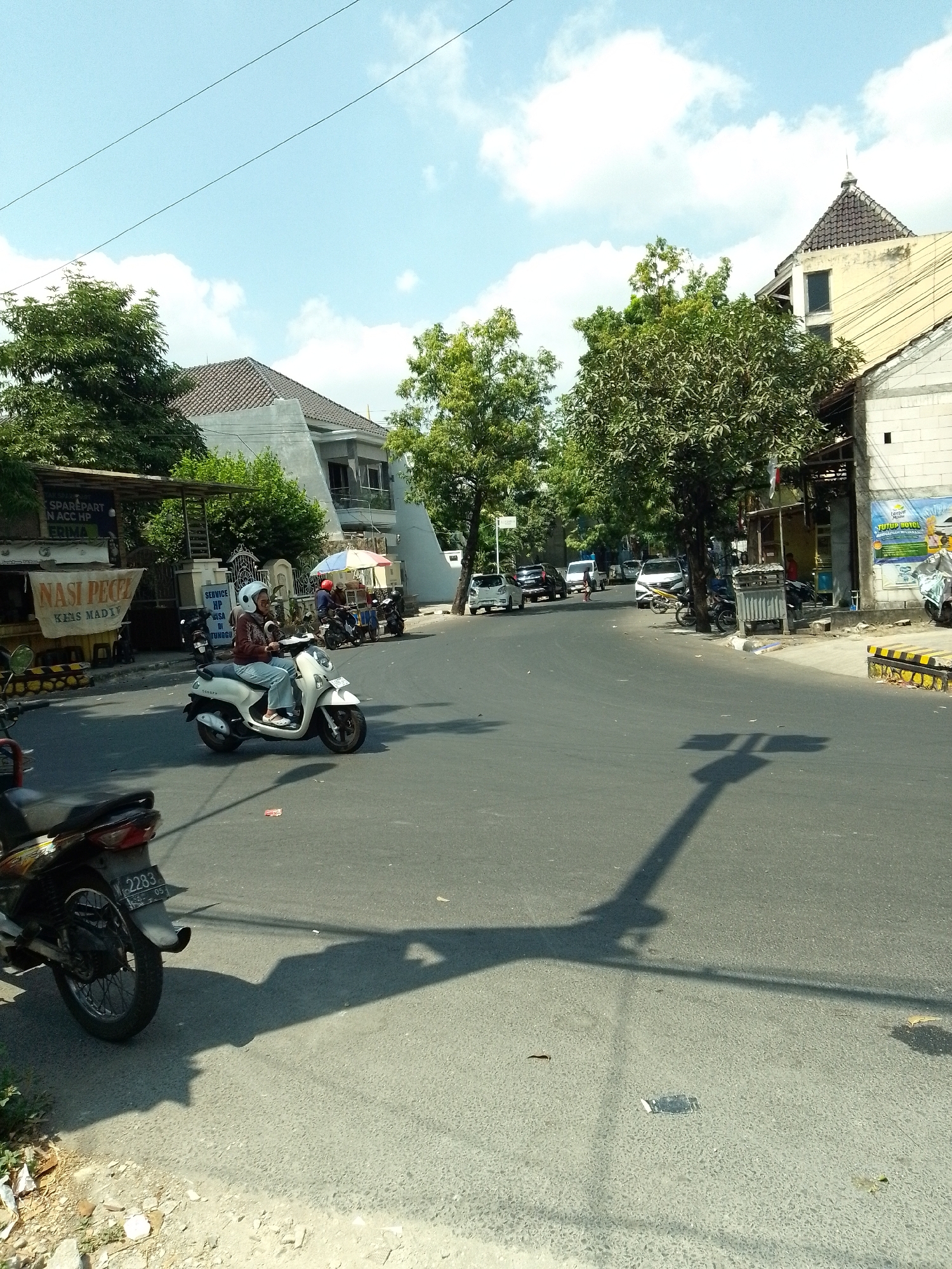 The image depicts a street scene in Tumenggungan, Indonesia, on a sunny day with scattered clouds. A woman wearing a helmet and a maroon long-sleeved shirt and blue jeans is riding a white scooter in the foreground, heading towards the left of the frame. Her scooter has the model name "SCOOPY" visible. To the left of the scooter, a portion of another dark-colored motorcycle is visible, parked on the side of the road.
The street is paved with asphalt. On the left side of the street, there is a food stall with a yellow banner that reads "NASI PECEL" and "KHAS MADIYUN." Above the food stall, a sign reads "SPAREPART N ACC HP ERIMA." Behind this stall, there is a building with a grey tiled roof and white walls. Further down the street, several other vehicles are visible, including cars and motorcycles. One car, a white hatchback, is turning the corner to the left.
On the right side of the street, there are several buildings. One building has a light yellow facade and a dark grey tiled pyramid-shaped roof. Another building to its right is constructed with white bricks. A few trees with green foliage are scattered throughout the scene, providing shade. The shadows of trees and a pole are cast across the road. In the distance, more buildings and trees are visible, indicating a moderately urban environment. A white car is seen driving away from the camera on the right side of the street.