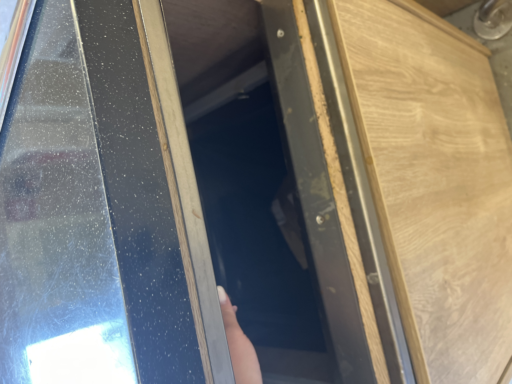 An image shows a person's hand reaching into a dark opening under a countertop in what appears to be a kitchen. The countertop is black with white speckles and has a metallic trim. The opening leads to a dark interior, possibly a cabinet or appliance. The lower part of the image shows a wooden cabinet with a visible grain pattern. There is no indication of the location in Volzhsky, Russia.