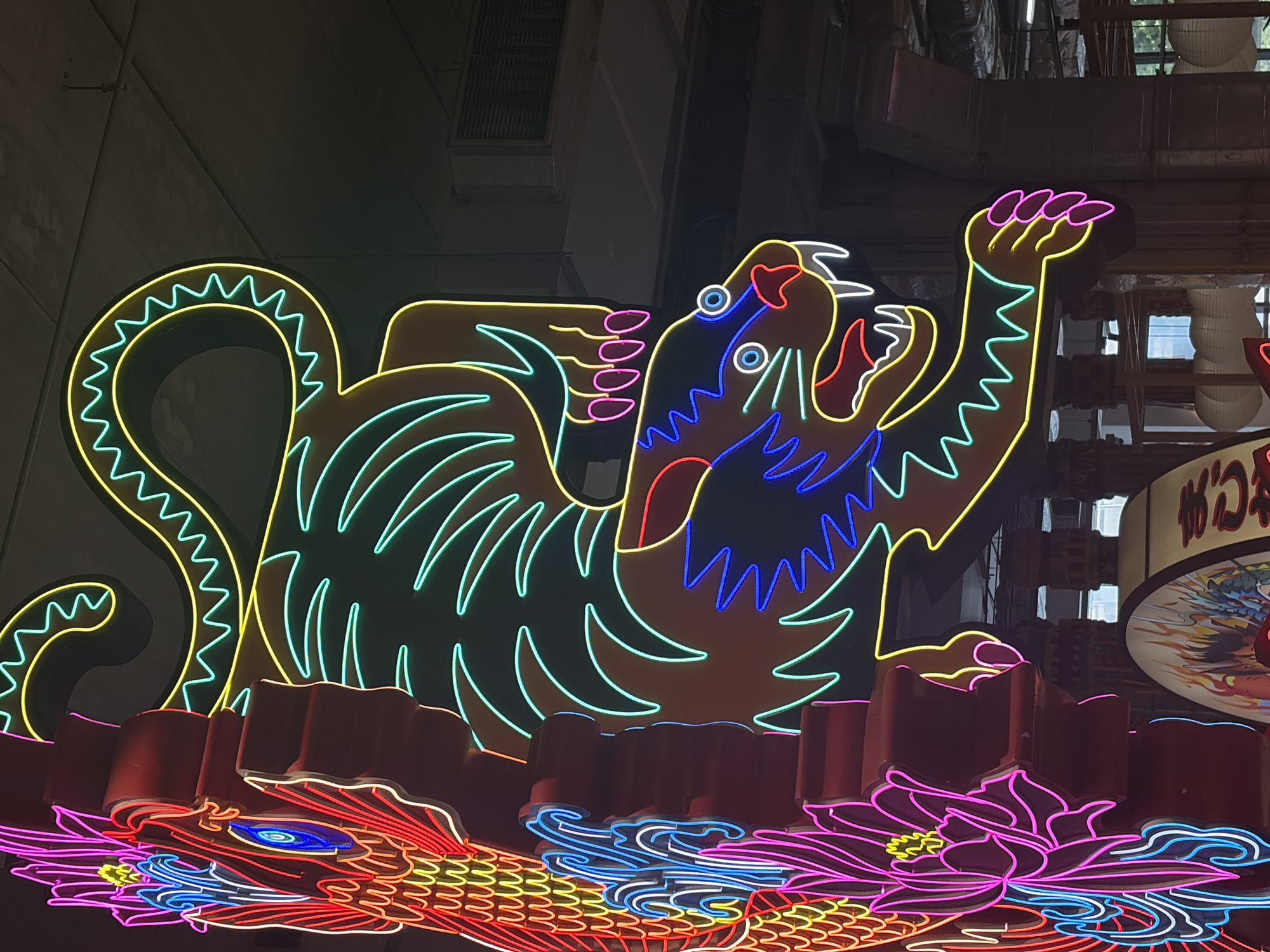 Here is a detailed description of the image:
The image shows a vibrant, neon-lit display in Bangkok, Thailand. The main focal point is a stylized tiger figure, outlined in bright yellow, blue, red, and pink neon lights. The tiger appears to be in a defensive or attacking posture, with its claws extended and mouth open.
Beneath the tiger, there's another neon display, possibly representing a dragon, with scales outlined in red, yellow, and blue. Interspersed with the dragon are stylized lotus flowers, outlined in pink neon. These are set against a background of dark colors, likely the building exteriors and nighttime sky. The scene suggests a lively, urban environment, typical of Bangkok's bustling street scenes. There are buildings in the background, and ventilation systems that are illuminated, although the focus is on the neon sign.