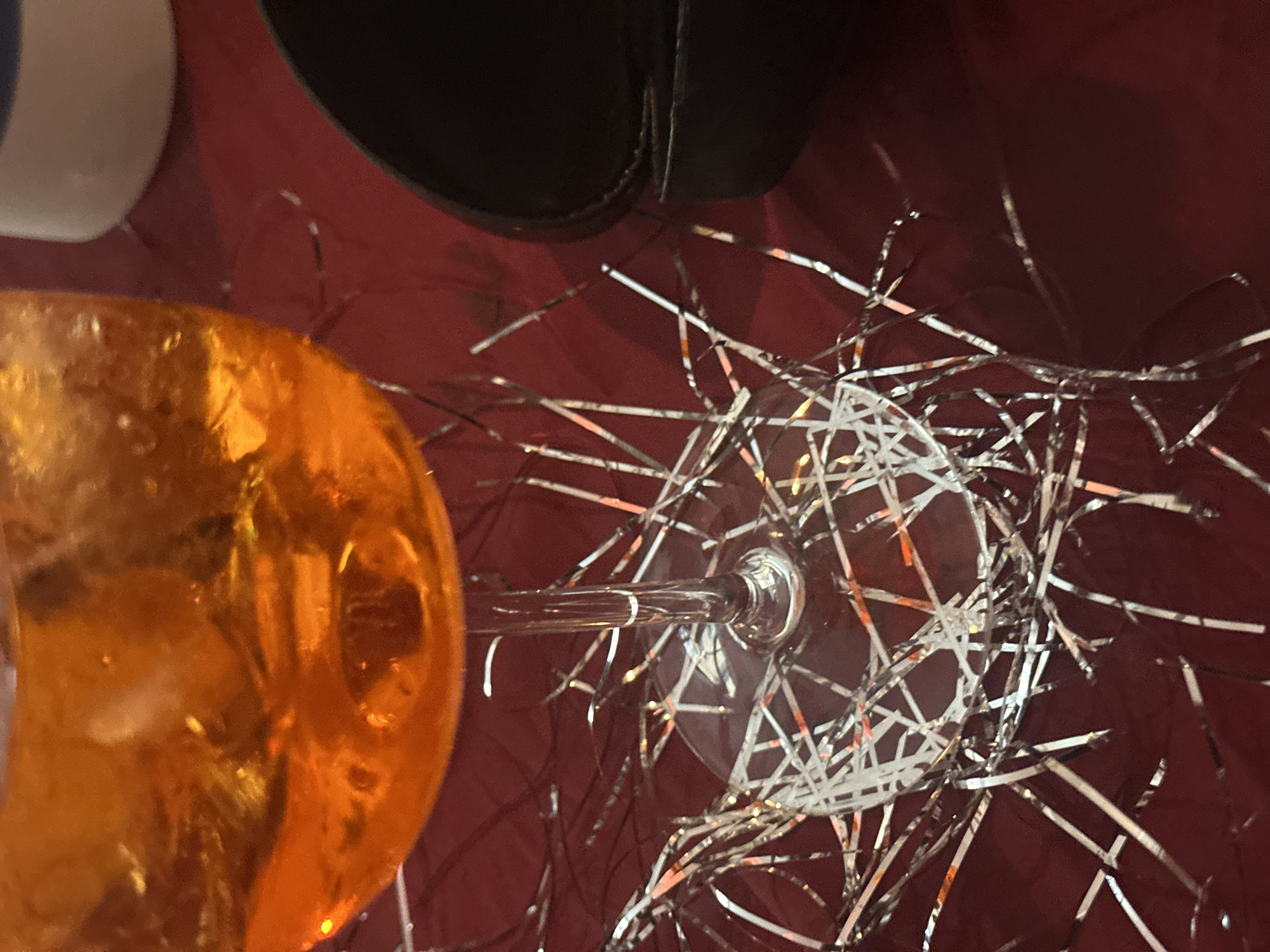 The image shows a close-up, overhead view of celebratory items on a dark red surface. A partially visible orange-colored glass is on the left. A black object, possibly a shoe, is partially visible at the top of the frame. There is a clear stemmed glass laying on its side. The scene is scattered with silver tinsel. The location and information regarding people or interactions is not available from the image.