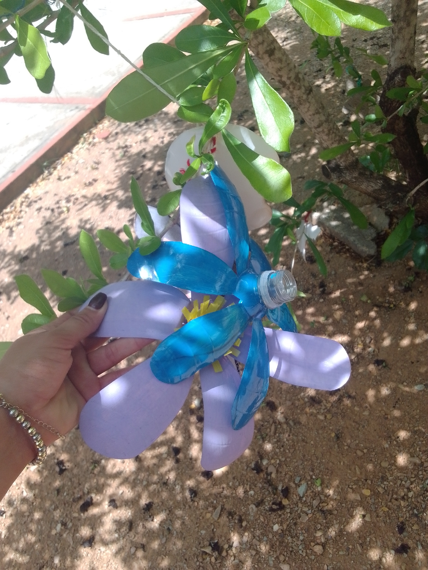 A hand with dark nail polish and a gold beaded bracelet is holding a homemade flowercraft. The flowercraft is made from a blue plastic bottle and purple petals. The petals are a light lavender color and are shaped like teardrops. The flower has a yellow center. The background of the image is a tree with green leaves and brown bark. The ground is covered in sand and small pebbles. The image appears to have been taken outdoors during the day. The light suggests it is a sunny day. The location cues suggest it is likely La Tagua, Venezuela, a tropical or subtropical region with sandy ground and lush vegetation. There is no visible text in the image.