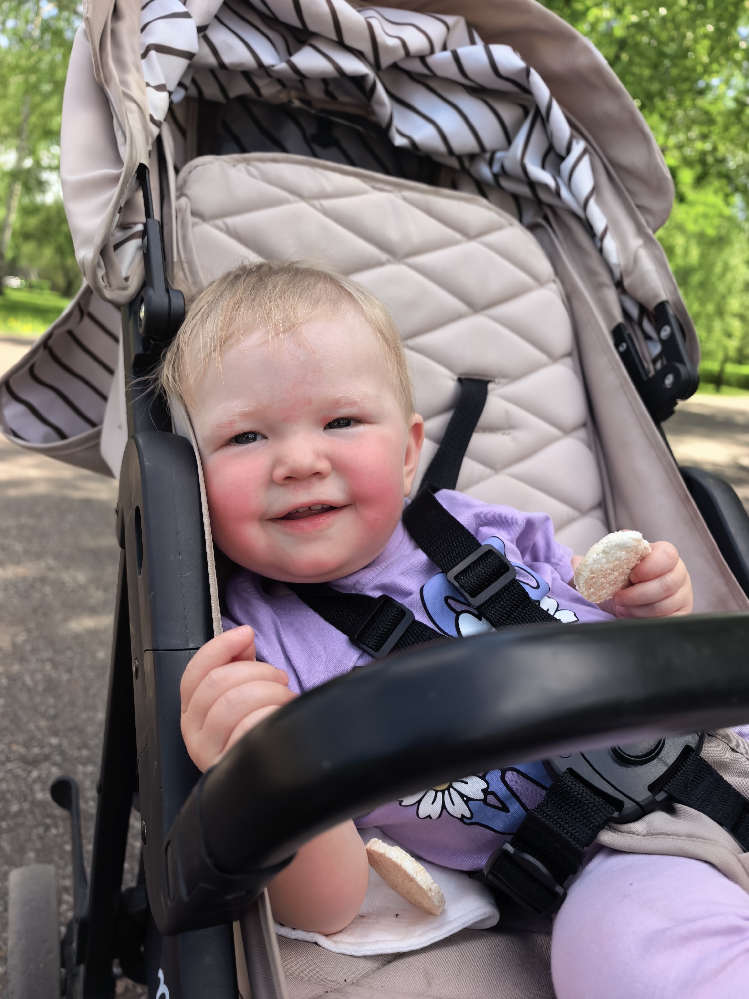 A baby with blonde hair and rosy cheeks sits in a stroller outdoors, holding a small, round snack. The baby wears a purple shirt with a daisy graphic and is secured with black straps. The stroller has a beige quilted interior and a striped canopy. The background shows greenery, suggesting a park or other green space. The location is the city of Sterlitamak, Russia. The surface on the left indicates a paved path.