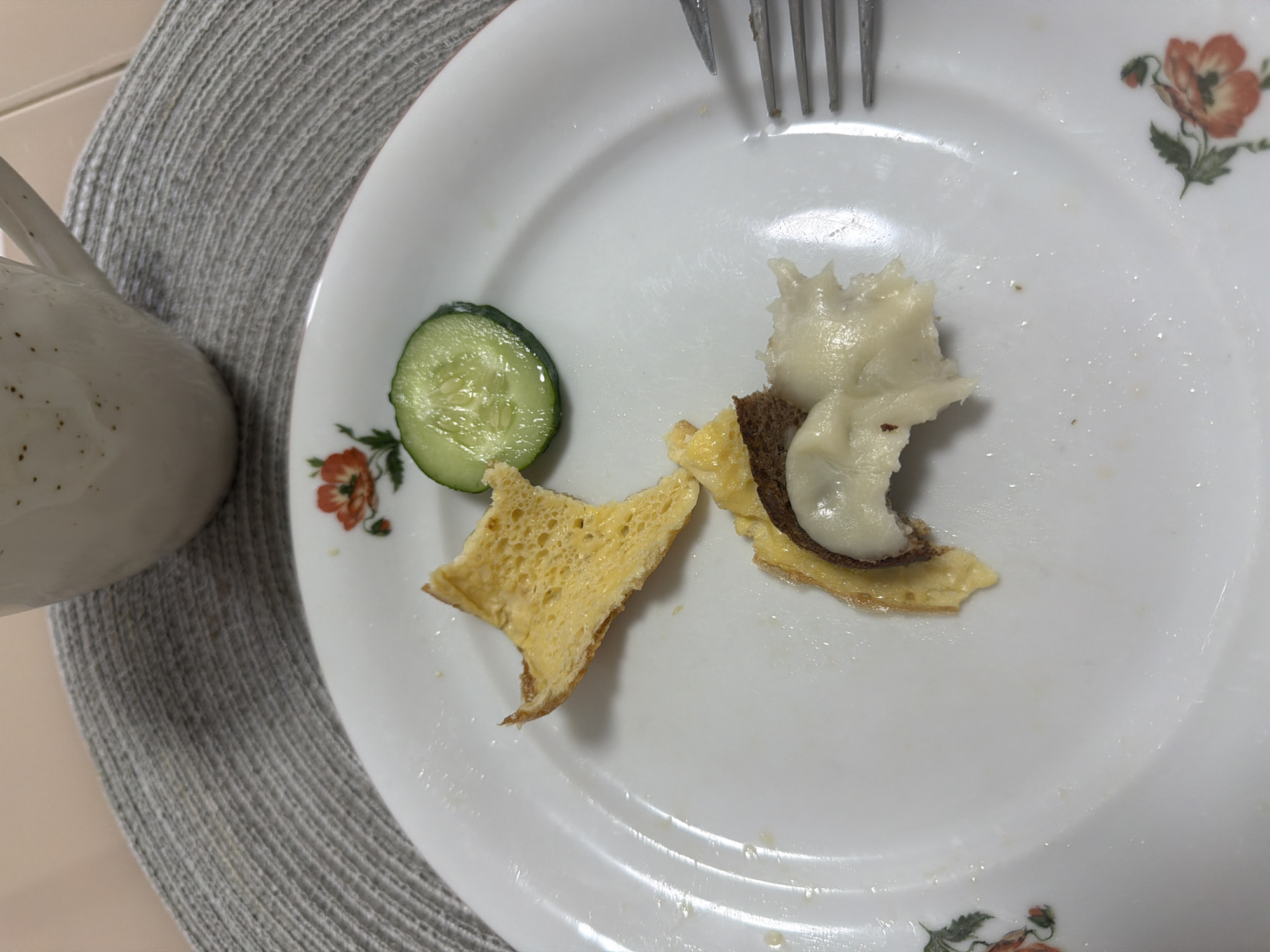 A white plate with a floral pattern contains remnants of a meal consisting of a cucumber slice, a piece of omelet, and a small sandwich with brown bread, what appears to be a fried egg and cheese/sour cream on top. A fork is resting on the plate. A mug and a grey placemat are visible to the left of the plate. The location and time are not provided in the original image, thus I cannot fulfill that request.