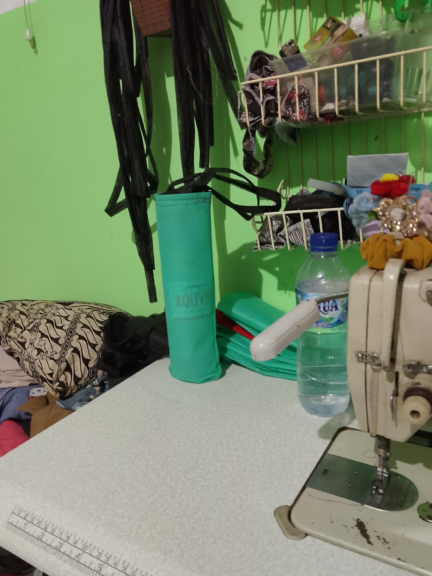 In the city of Kelurahan, Indonesia, this image captures a cluttered domestic workspace, likely a sewing room. The scene is dominated by a light green wall and a light-colored tabletop, hinting at a room that may not receive abundant natural light.
On the left, a partially visible patterned textile, possibly a batik cushion, rests against other fabrics. To its right, a tall, teal cylindrical case with "AQUVE" printed vertically stands upright. Behind this, several black straps or belts hang from above, casting shadows on the wall.
Further right, a wire rack is attached to the wall, holding various items. The upper shelf contains a clear plastic container filled with fabric scraps, buttons, and other crafting materials. Below this, another shelf holds more assorted items, including colorful hair accessories. A clear plastic bottle of "AQUA" mineral water sits on the tabletop, its blue cap visible. A white plunger-like object is positioned near the bottle, potentially a sewing machine accessory or a household item.
In the foreground, a beige sewing machine is partially visible, its needle and presser foot in focus. The tabletop itself is patterned and features a ruler graphic embedded along its edge, indicating it's a dedicated workspace. The overall impression is one of a functional, albeit disorganized, creative environment.
