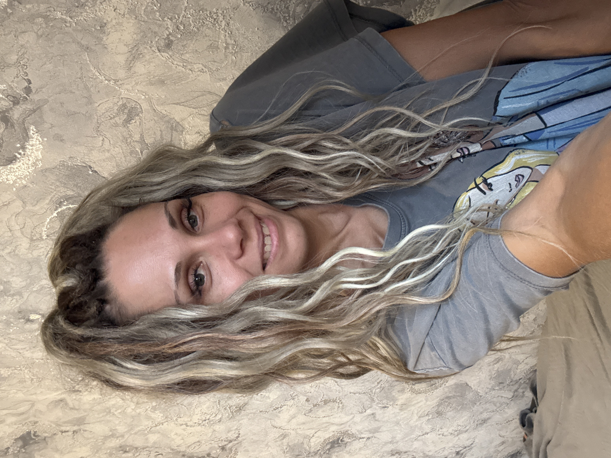 A woman with blonde and brown dreadlocks, styled in a high ponytail, smiles at the camera. She is wearing a grey t-shirt with a graphic print of two women. Her eyes are green and she has on dramatic false eyelashes. The background is a textured, beige and gold wall. The image appears to be a selfie taken indoors, possibly in a bedroom or living area.
