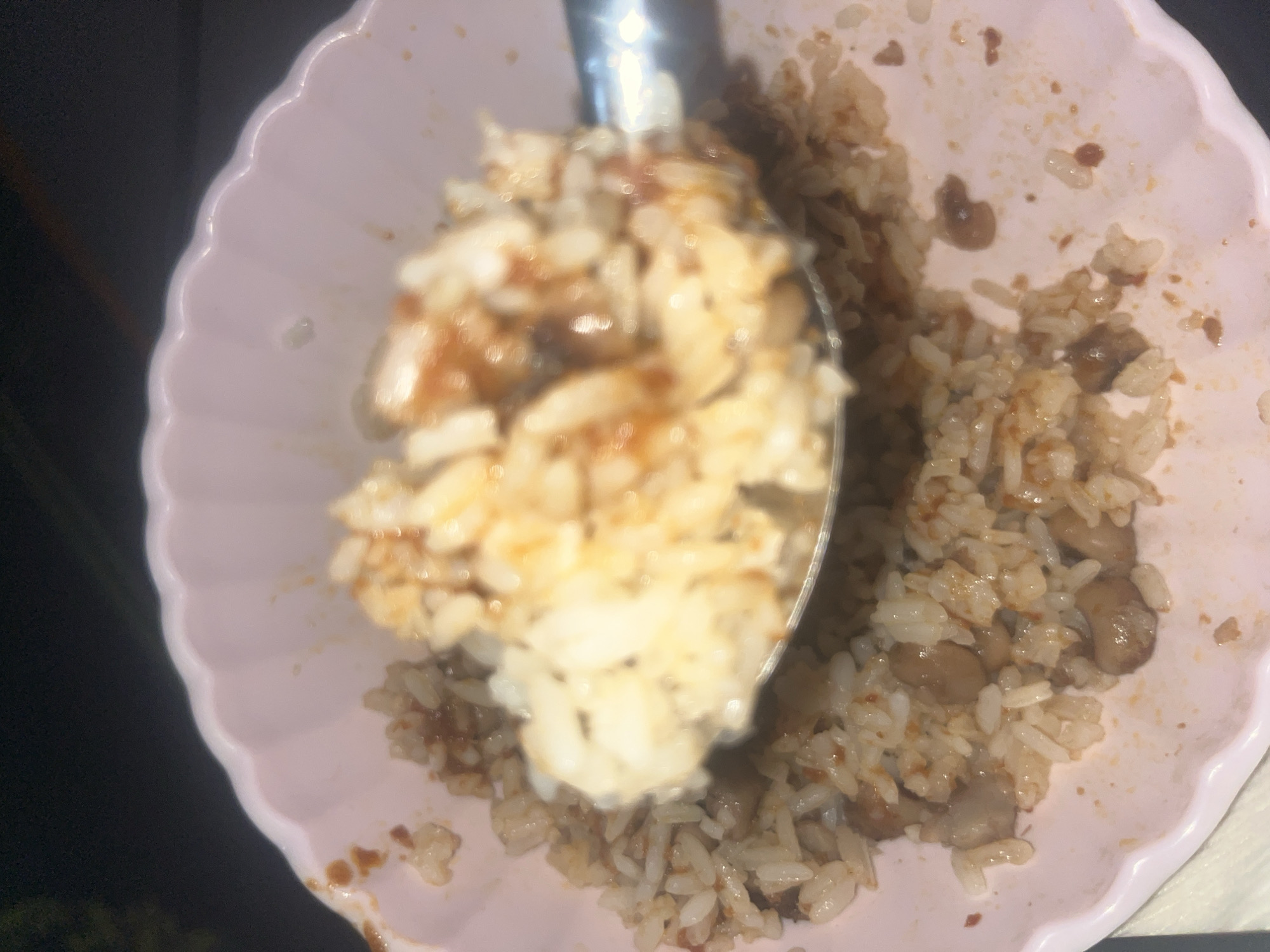 A close-up shot shows a light pink scalloped bowl filled with rice mixed with a brown sauce and beans, possibly lentils. A silver serving spoon scoops up a portion of the rice mixture. The scene appears to be indoors. Gura Top, Nigeria is not discernible in this image.
