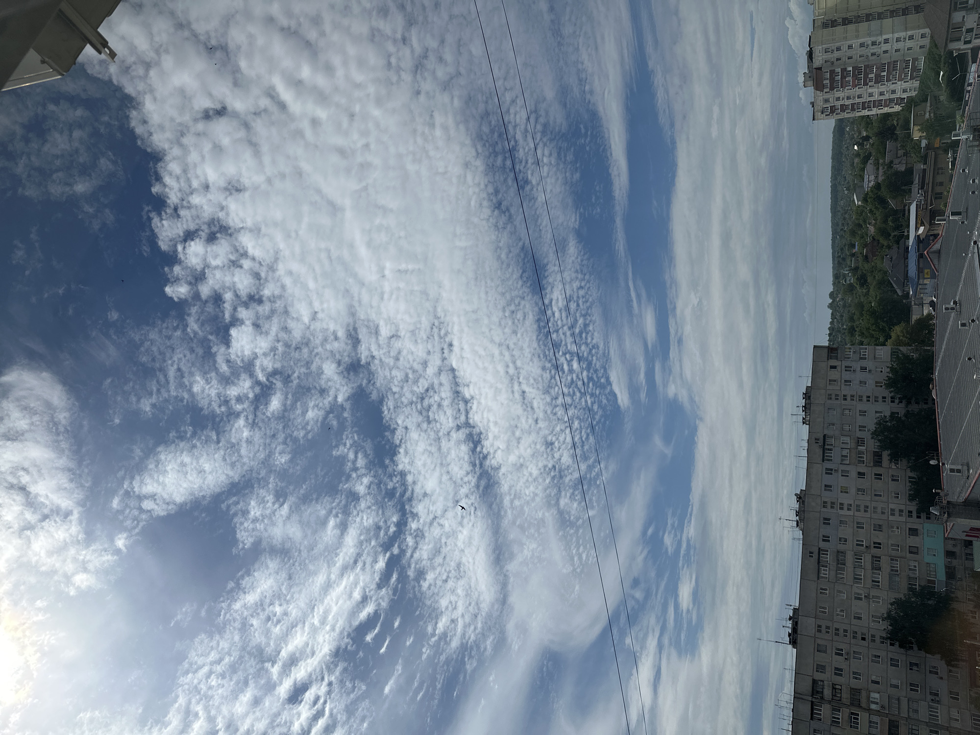 The image displays an urban landscape under a bright, partly cloudy sky, captured from an elevated perspective, likely a building or balcony. The scene is oriented horizontally, with the sky dominating the upper two-thirds of the frame and the cityscape occupying the bottom third.
The sky features a mix of blue patches and extensive white clouds. Towards the bottom-left, the sky is clearer blue, with a bright, hazy glow indicating the position of the sun. Moving upwards and to the right, the clouds become more prominent, appearing as textured, often rippled or streaky formations, suggestive of altocumulus or cirrocumulus clouds, stretching across the majority of the visible sky. Several small, dark silhouettes of birds are visible against the bright sky, particularly in the left and center portions. Two thin, dark, parallel utility lines or power cables diagonally traverse the sky from the bottom-left towards the top-right.
The urban landscape at the bottom of the image consists primarily of multi-story residential buildings. On the right side, a tall, light-colored apartment building with numerous windows and balconies is prominent. Extending across the lower part of the image, other residential blocks, appearing somewhat older with greyish facades, are tightly packed. Patches of green trees and foliage are visible between and behind the buildings, extending towards the distant horizon, which appears hazy. A dark roof, possibly metal, is visible in the bottom-right corner.
The time of day is daytime, likely a bright morning or afternoon, given the clear illumination. The weather is partly cloudy. No discernible text is visible anywhere in the image.