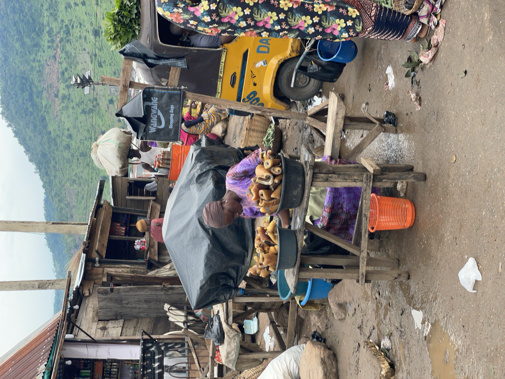 The image depicts a bustling outdoor market scene in Lokoja, Nigeria. In the foreground, a woman is tending to her stall, which is laden with a variety of food items, possibly fried snacks or pastries, displayed in large dark buckets. She is wearing a purple patterned dress and a brown headscarf. Behind her, another woman in a colorful floral dress is partially visible.
The market appears to be situated on a dirt road, with puddles of water suggesting recent rainfall or a wet environment. A yellow motorized tricycle, likely used for transport or as part of the market stall, is parked nearby. It has visible text on its side, including "DA" and "900". In the background, a lush green hillside rises, indicating a location with natural beauty.
Various objects are scattered around the market, including a large black tarp covering some goods, a bright orange plastic basket, blue buckets, and discarded plastic bags. Wooden structures, forming stalls and shelters, are visible throughout the scene. The overall impression is one of a vibrant, active marketplace, likely operating during the daytime under overcast or humid weather conditions. The scene conveys a sense of everyday commerce and community life.