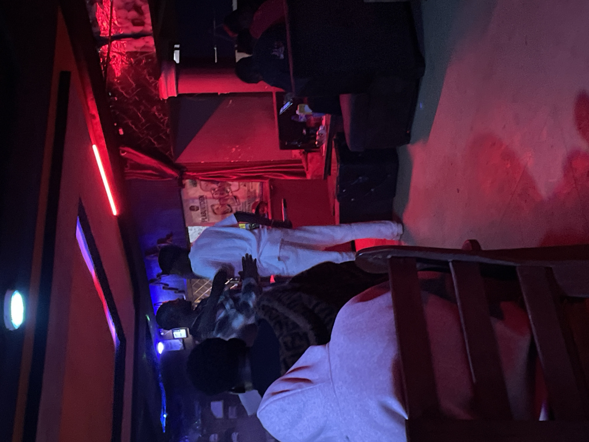 The image depicts an indoor scene, likely a bar or club, with predominantly red and blue lighting. In the foreground, a person wearing a light pink hooded sweatshirt is seated, facing away from the camera, their back to a wooden chair. To their left, another individual, also facing away, is wearing a patterned top. Further back, two men are interacting. One, wearing a plaid shirt, appears to be gesturing, while the other, in a white t-shirt and pants, is facing away from the camera. In the background, a poster with text is visible on the wall, and a table with bottles and a phone is present. Several dark seating arrangements are also visible. The overall atmosphere suggests a social gathering in a dimly lit entertainment venue. The location is identified as Durumi, Nigeria.